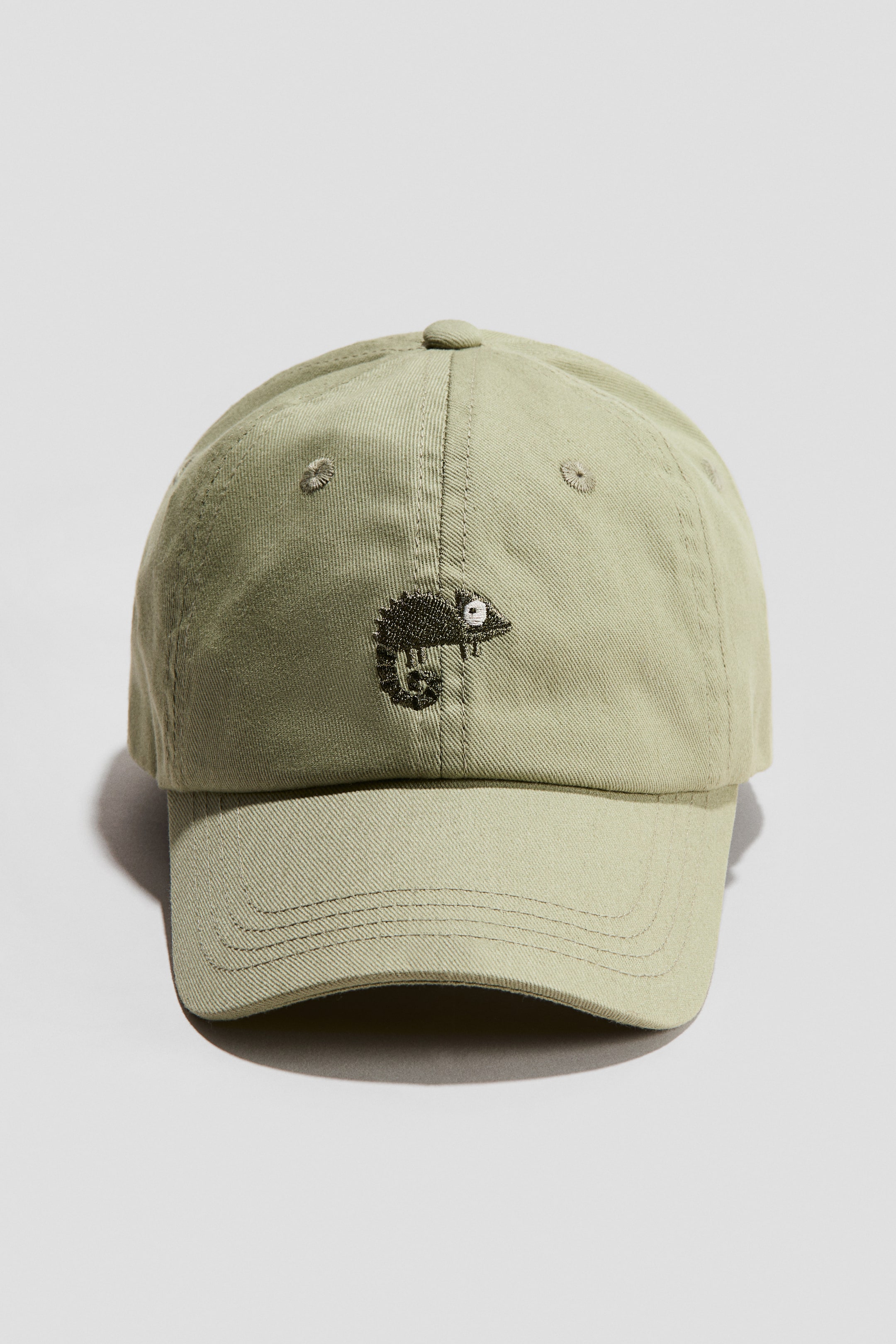 Agrandir l'image: Une casquette vert olive clair est présentée de face, fabriquée en sergé de coton. Elle arbore un motif de caméléon brodé en vert foncé sur le devant, ainsi que des œillets brodés, et une languette ajustable avec une fermeture à crochets et boucles à l'arrière.