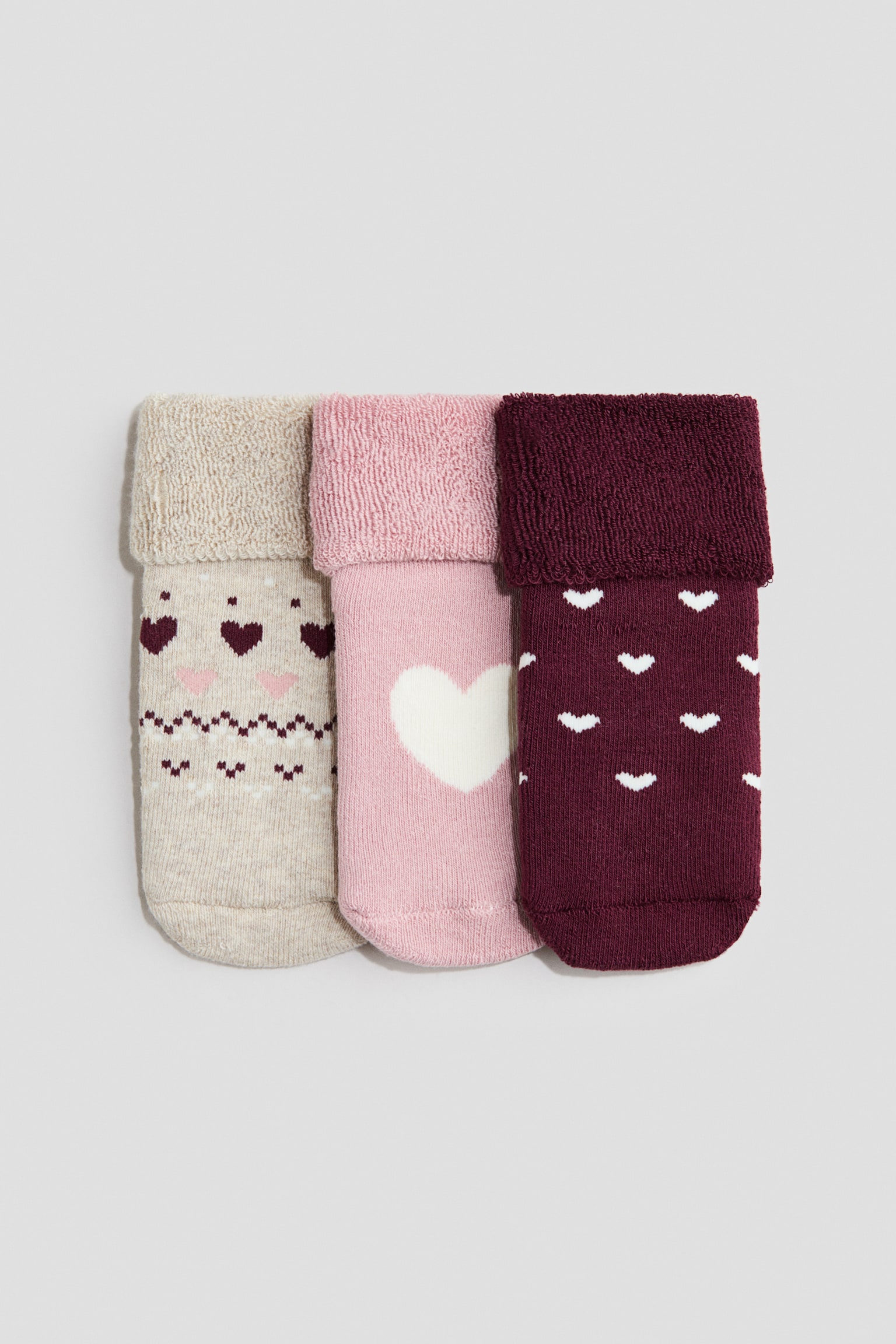 Tre stickade strumpor ligger bredvid varandra med framsidan vänd mot kameran och visar nedvikta skaft med ett strukturerat foder i frottéstickning. Urvalet شاÙ
Ùáº? includes a beige sock with a Fair Isle-inspirerad hjärtÂmönster, en ljusrosa strumpa med ett enda stort vitt hjärta och en vinröd strumpa med smÃ¥ vita hjärtan.