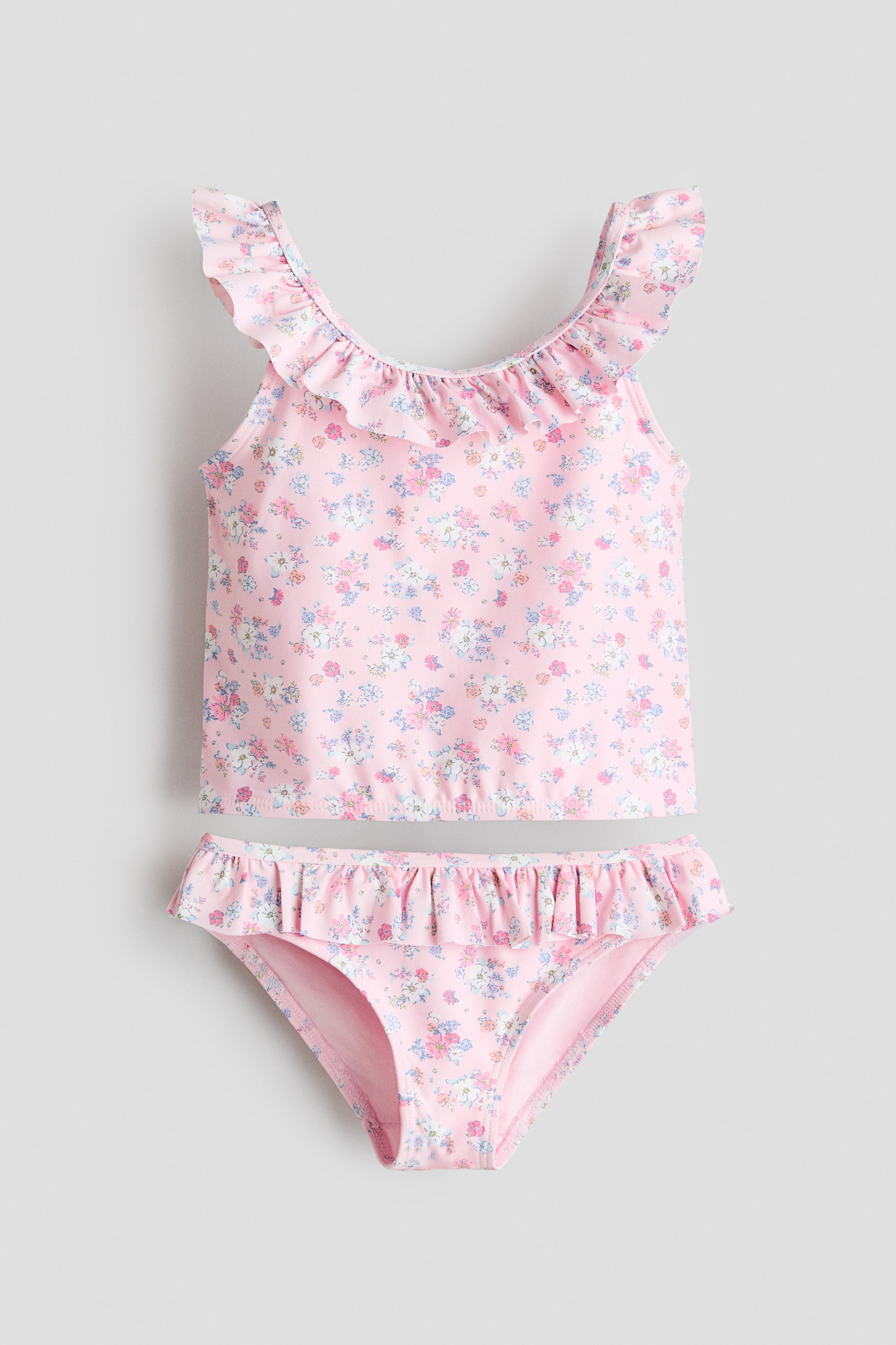 Visa större bild: Ett rosa tankiniset med blommönster visas FRONT, med en överdel med smala axelband och en volangkant som fortsätter från framsidan, över axlarna och runt halsringningen. Matchande underdelar visar en volangkant i midjan och ett fodrat grenparti.