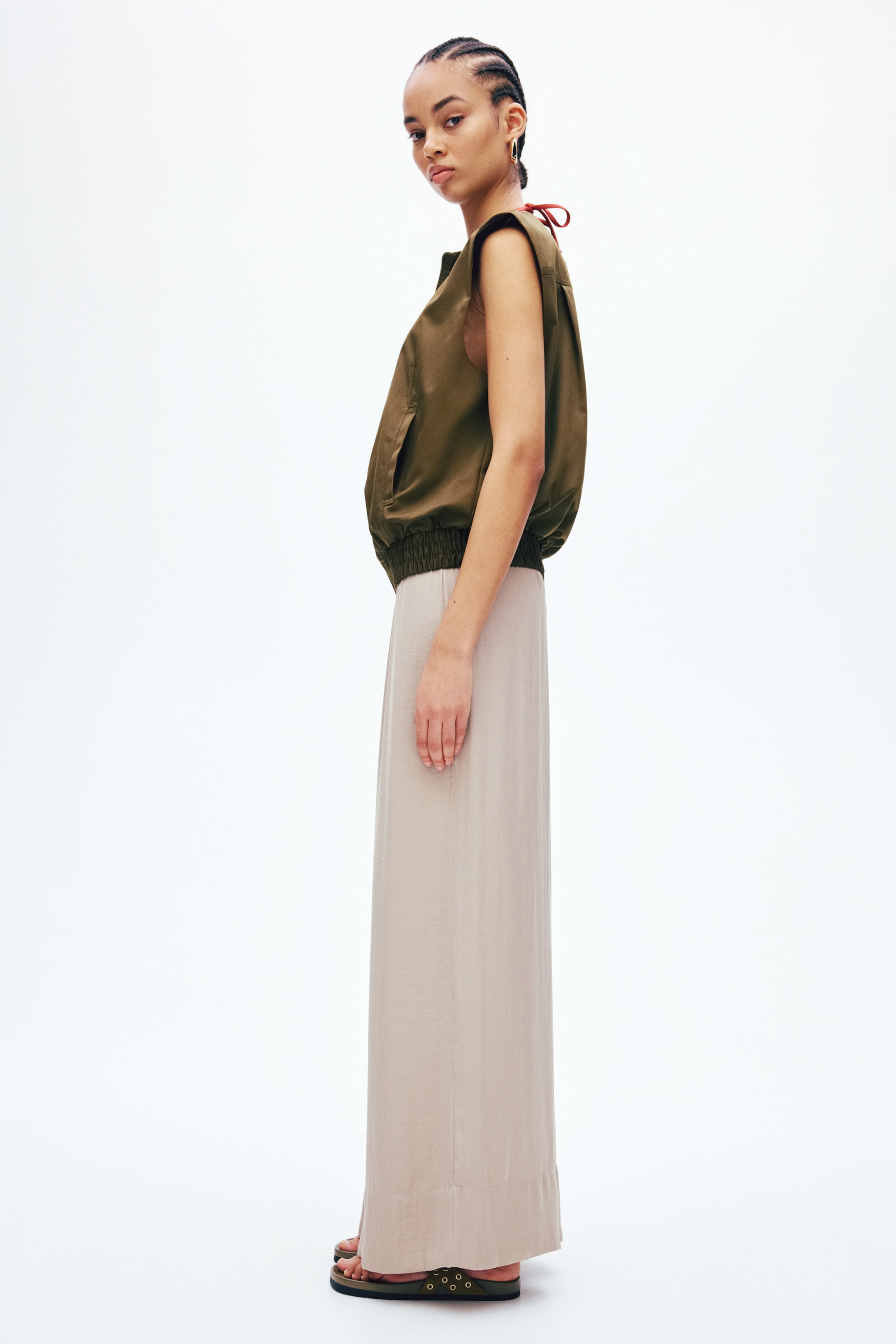 Voir une image plus grande: Un haut sans manches de style bomber vert olive, avec un ourlet côtelé et des poches latérales, est assorti à une longue jupe maxi beige clair. Le haut présente également un lien brun-rouge à l'arrière du cou, tandis que la jupe tombe jusqu'au sol, révélant des sandales à enfiler vert olive avec des œillets dorés.