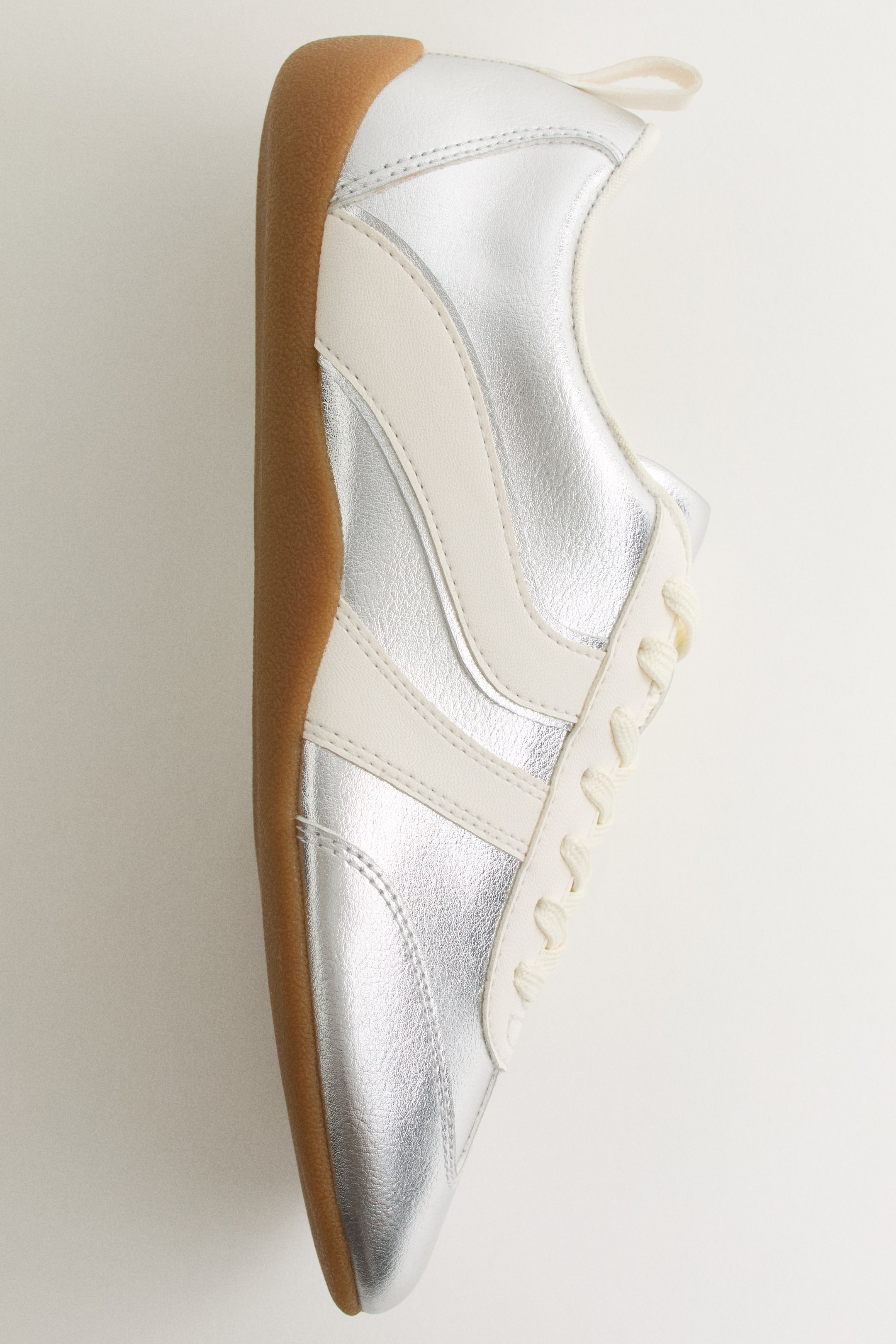 Größeres Bild ansehen: Ein Low-Top-Sneaker präsentiert seine rechte Seite und zeigt ein silbernes Metallic-Obermaterial mit cremefarbenen Lederstreifen und cremefarbenen Schnürsenkeln. Er verfügt außerdem über eine hellbraune Gummisohle und eine cremefarbene Zuglasche an der Ferse.