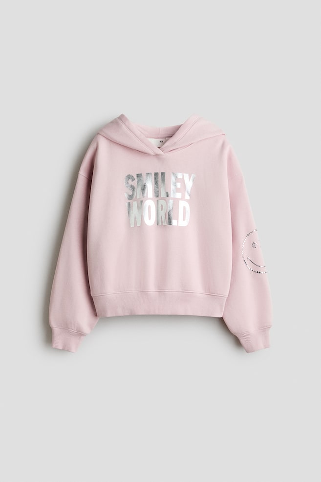 A pink hoodie showcases a metallic silver "SMILEY WORLD" graphic across its front. It also features a silver dotted smiley face design on its left sleeve and is completed with ribbed cuffs and a ribbed hem. - 1