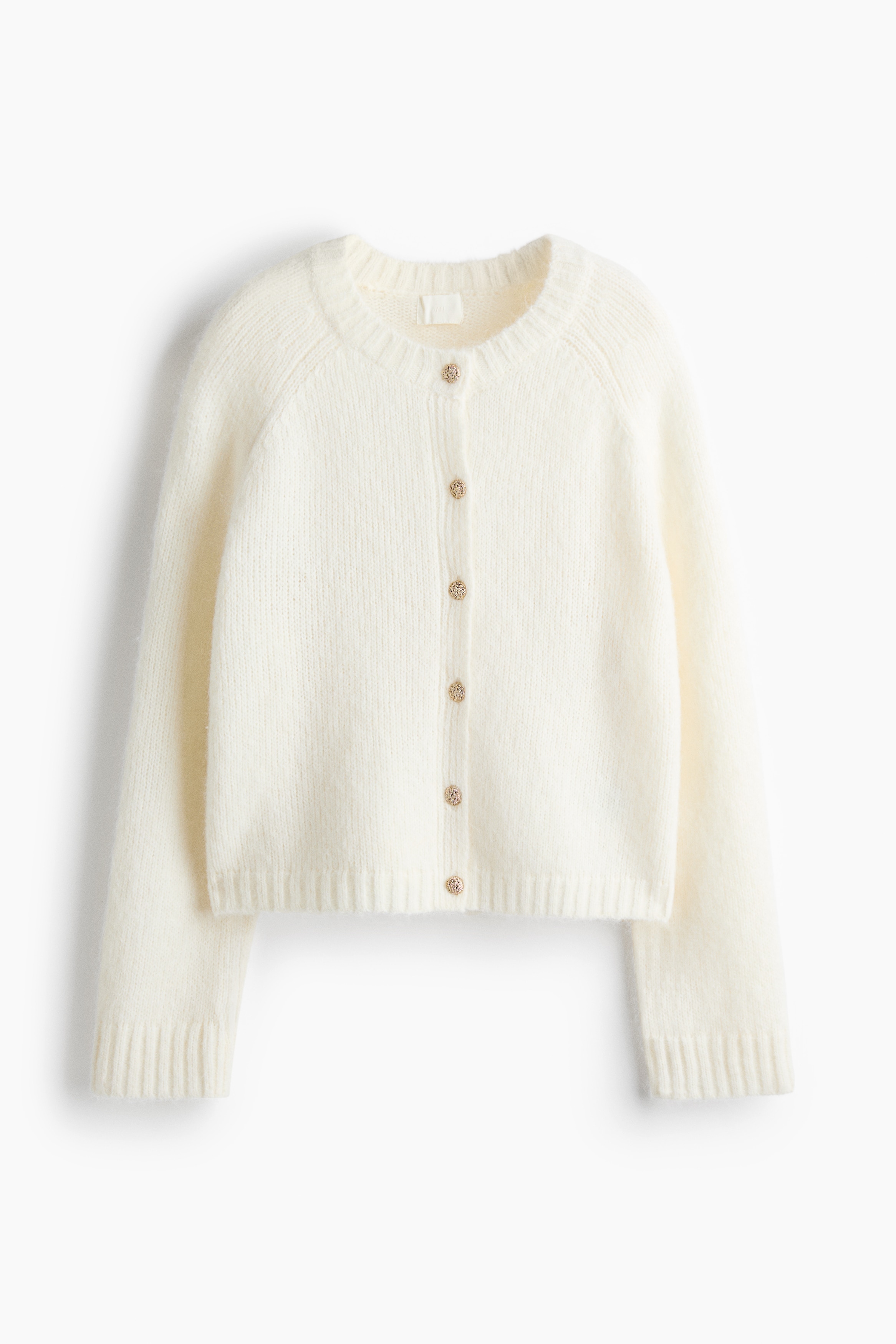 A short, oversized off-white knit cardigan featuring a round, rib-trimmed neckline, gold-tone buttons, and long raglan sleeves with ribbed cuffs is worn by the model. The model is standing with arms crossed, styling the cardigan with high-waisted white jeans.