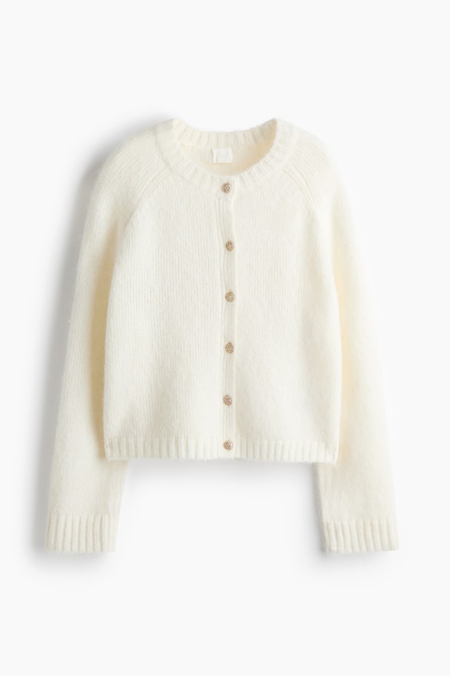 Eine cremeweiÃe Strickjacke ist von vorne zu sehen und präsentiert eine weiche, flauschige Textur sowie einen runden, gerippten Ausschnitt. Sie verfügt über einen vorderen Knopfverschluss mit sechs dekorativen Knöpfen in Goldoptik, lange Ãrmel mit gerippten Bündchen und einen gerippten Saum. - 1