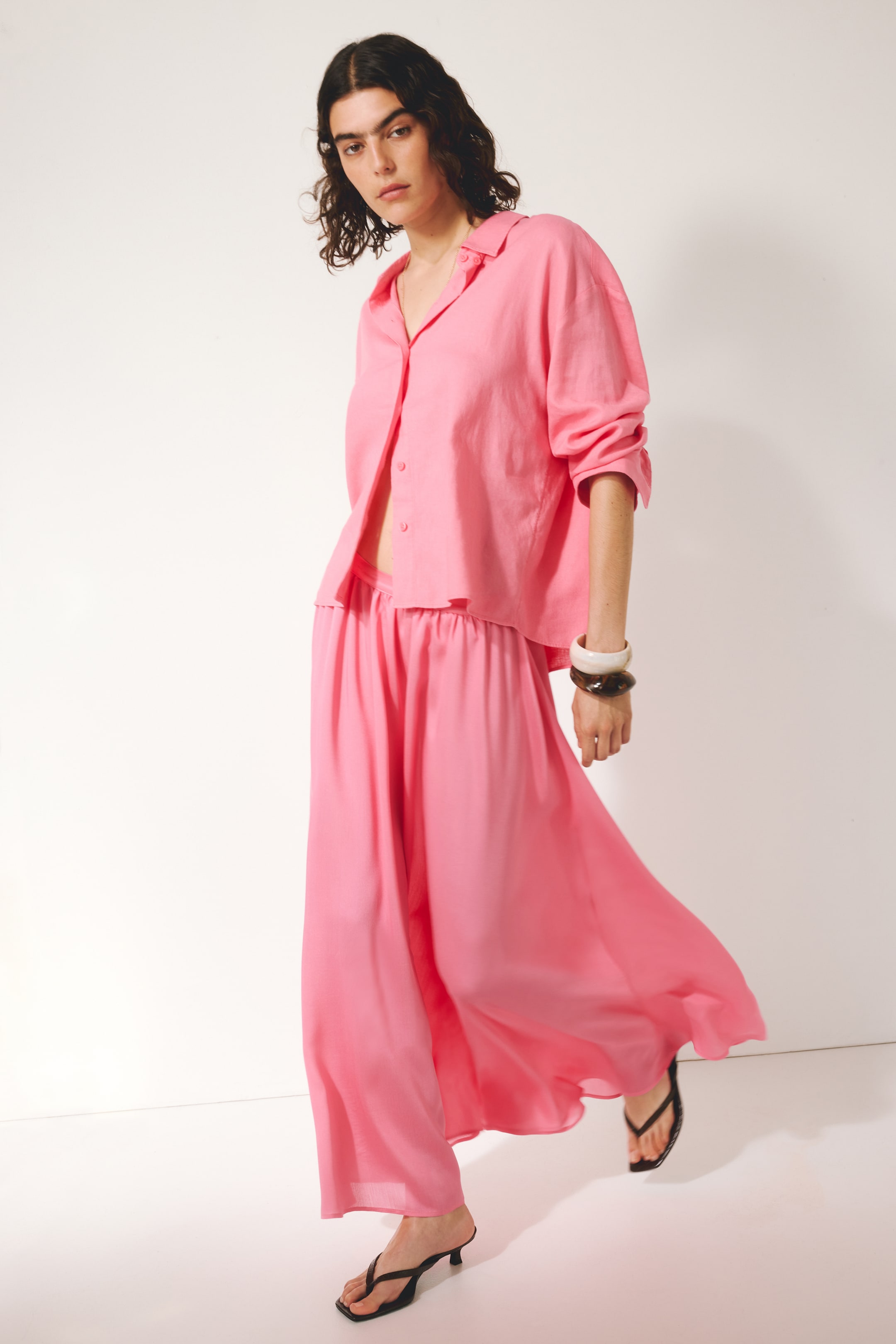 View larger image: The model is wearing a pink loose-fit skirt with a high-waisted design and gathered front. She rests a hand on the skirt's gathered waistband, while a white ribbed tank top is tucked into the skirt.