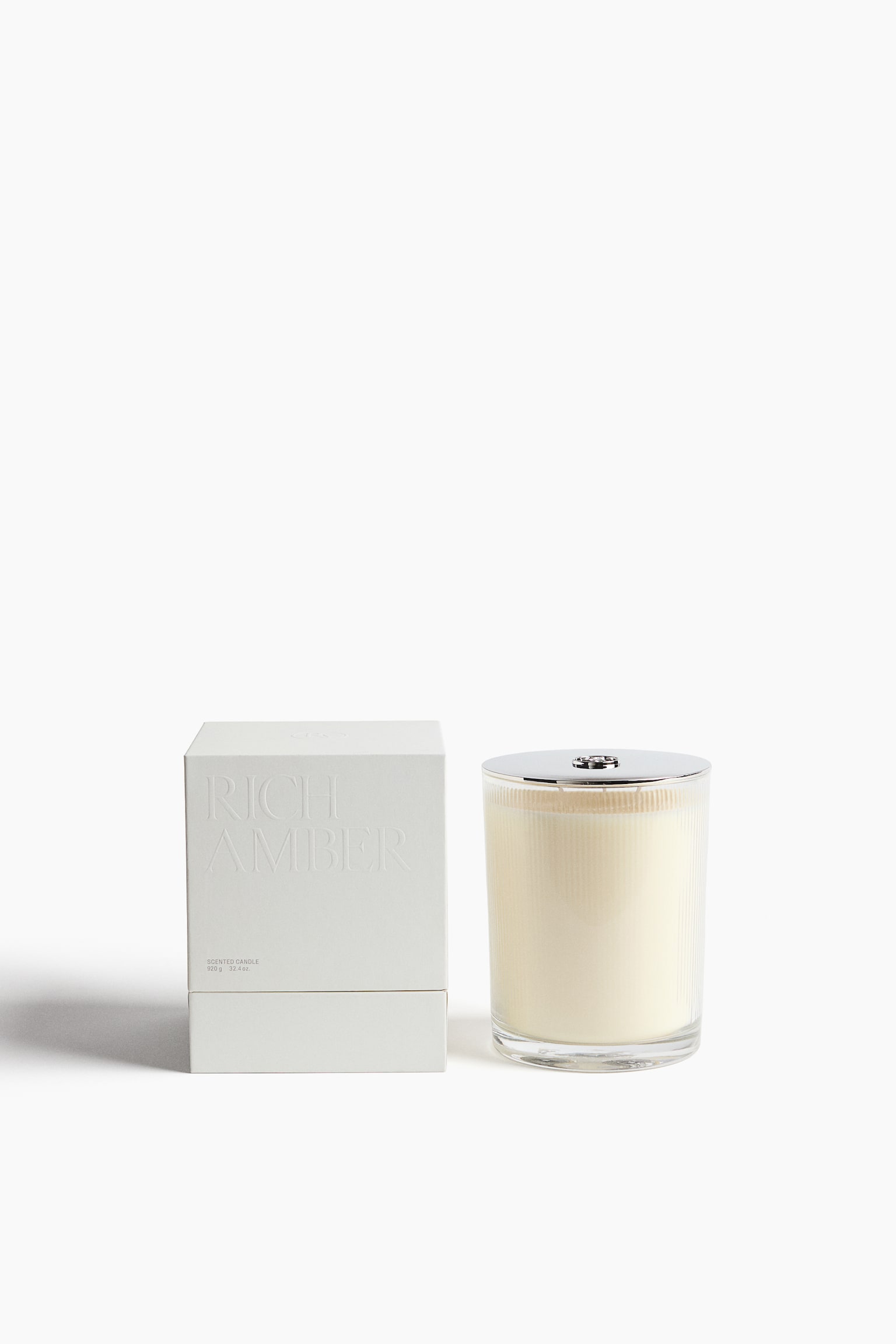Une bougie parfumÃ©e dans un bocal en verre transparent cÃ´telÃ©, rempli de cire de couleur crÃ¨me et surmontÃ© dâun couvercle argentÃ© poli, est placÃ©e Ã  cÃ´tÃ© de son coffret dâemballage blanc minimaliste. La face avant du coffret prÃ©sente le texte en relief Â« RICH AMBER Â» dans sa partie supÃ©rieure. - 1