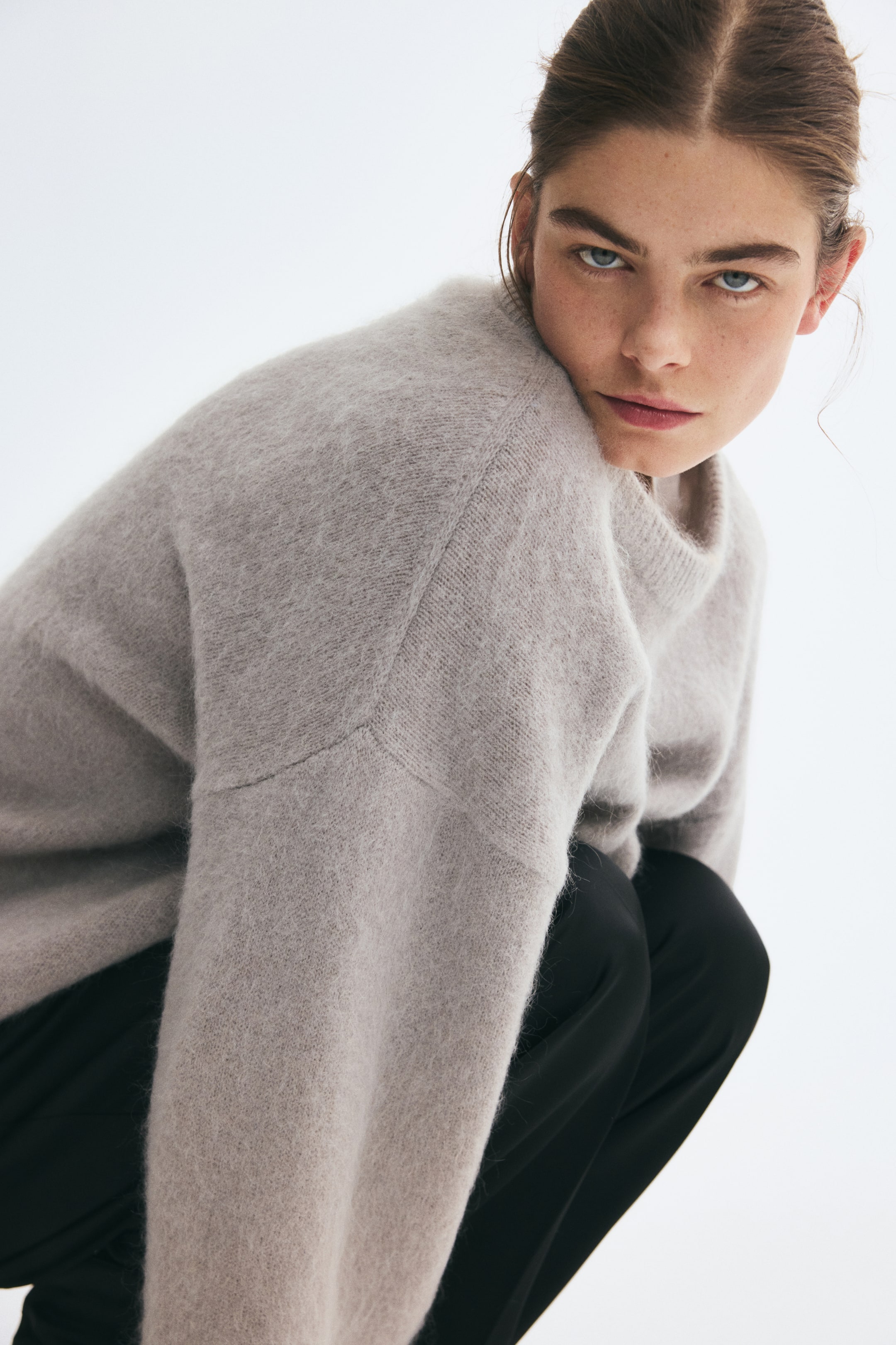 View larger image: An oversized, light grey knit sweater with a soft, fuzzy texture, dropped shoulders, and a ribbed crew neckline is worn by the model. The model is crouching while styling the sweater with black trousers.