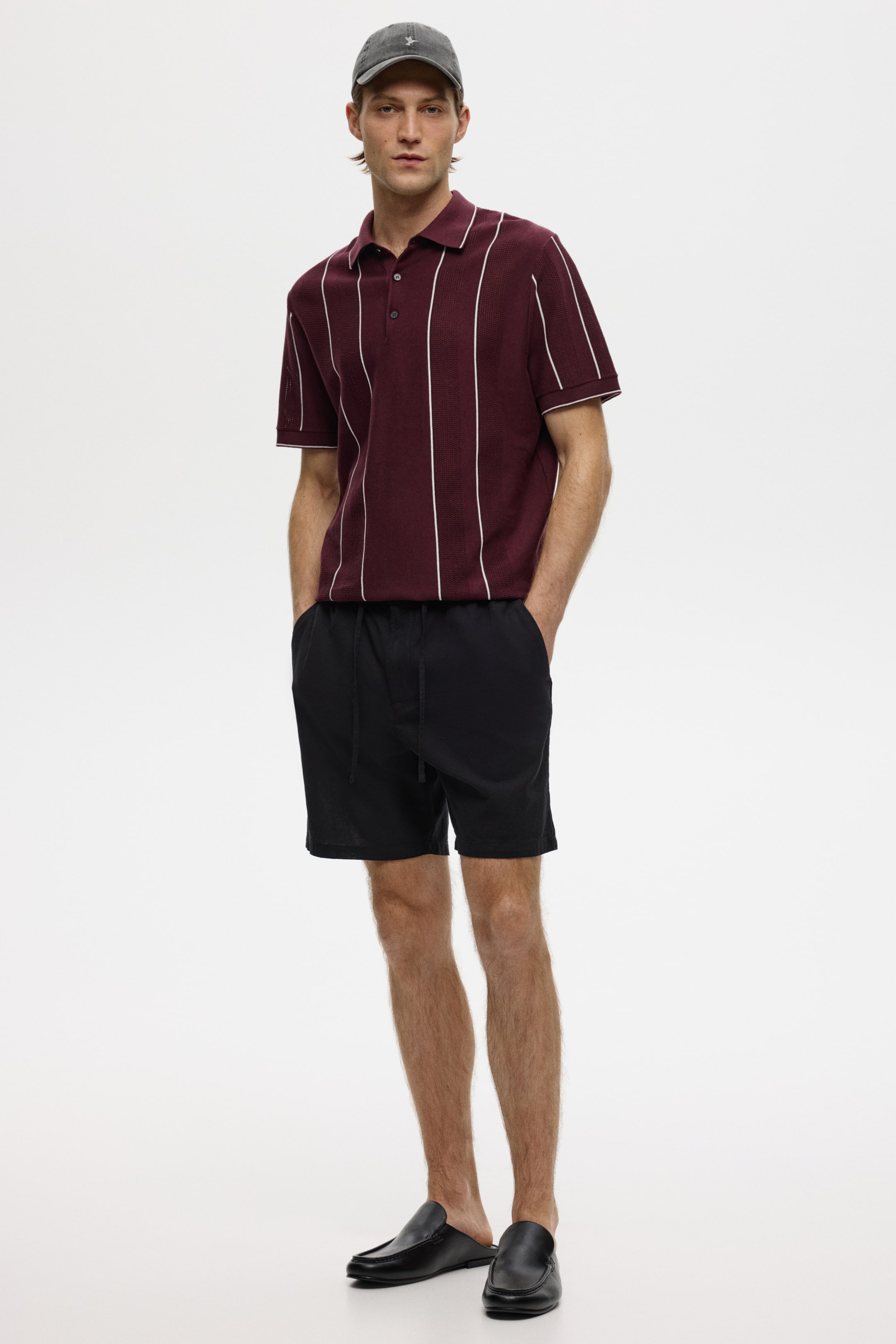 View larger image: A burgundy polo shirt with white vertical stripes, a two-button placket and white trim on the collar and short sleeves is being worn. It is paired with black shorts with an elasticated drawstring waistband and side pockets, falling to just above the knee.