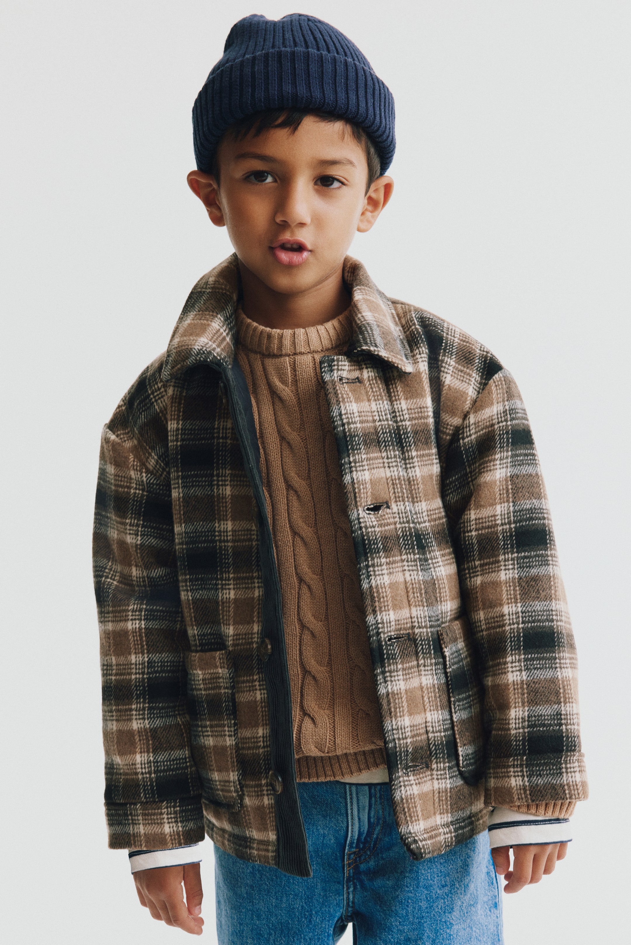 View larger image: A plaid padded jacket in a brown and beige brushed weave is worn open in a front view, layered over a brown cable-knit sweater. The jacket features a spread collar, dropped shoulders, open front pockets, and cuffed sleeves.
