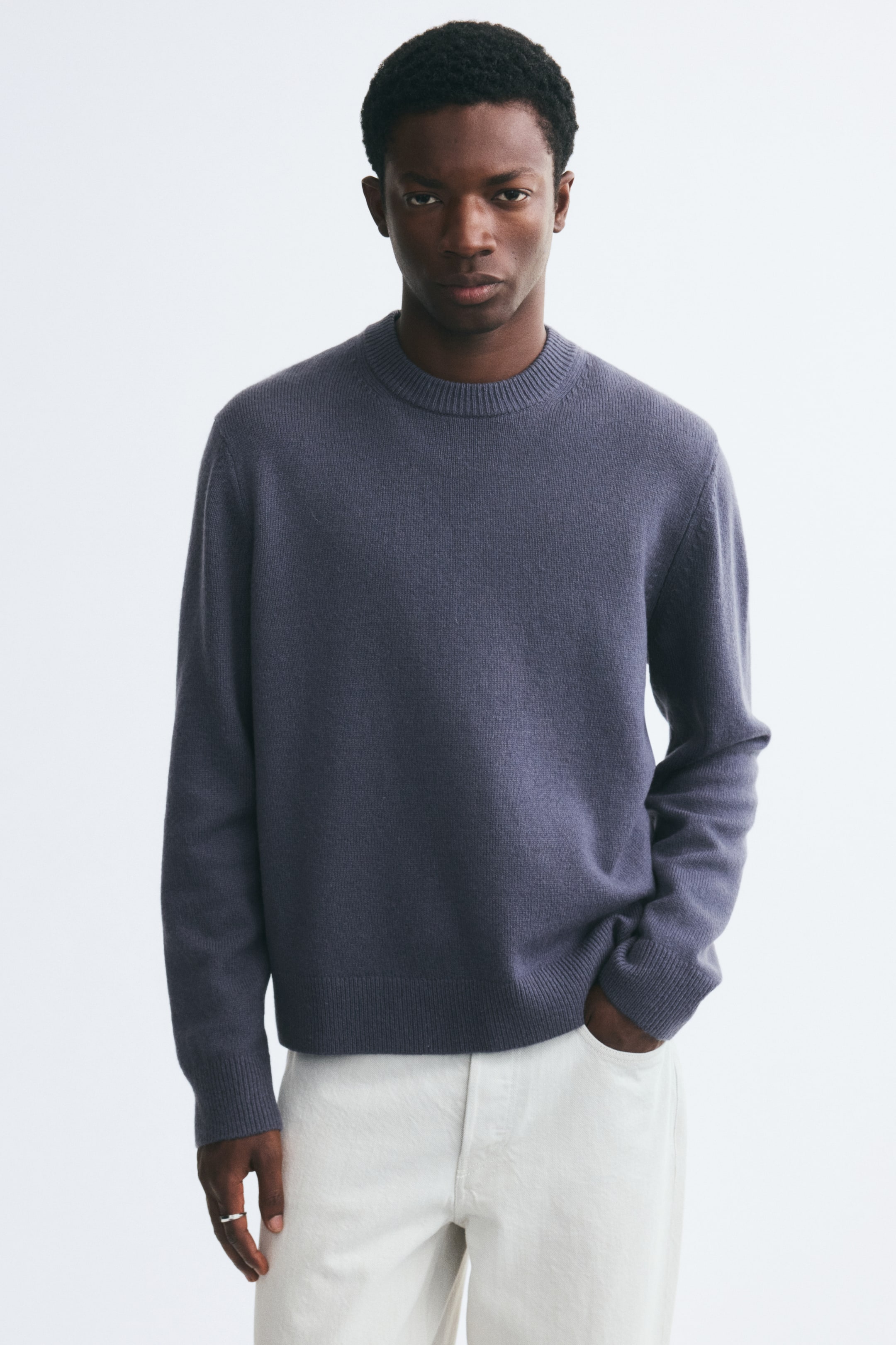 Größeres Bild anzeigen: Ein schieferblauer Wollpullover aus Strick mit rundem, geripptem Ausschnitt, langen Ärmeln und geripptem Saum wird vom Model getragen. Der Pullover in Regular Fit ist mit cremefarbenen Hosen kombiniert, wobei das Model eine Hand in der Tasche ruhen lässt.