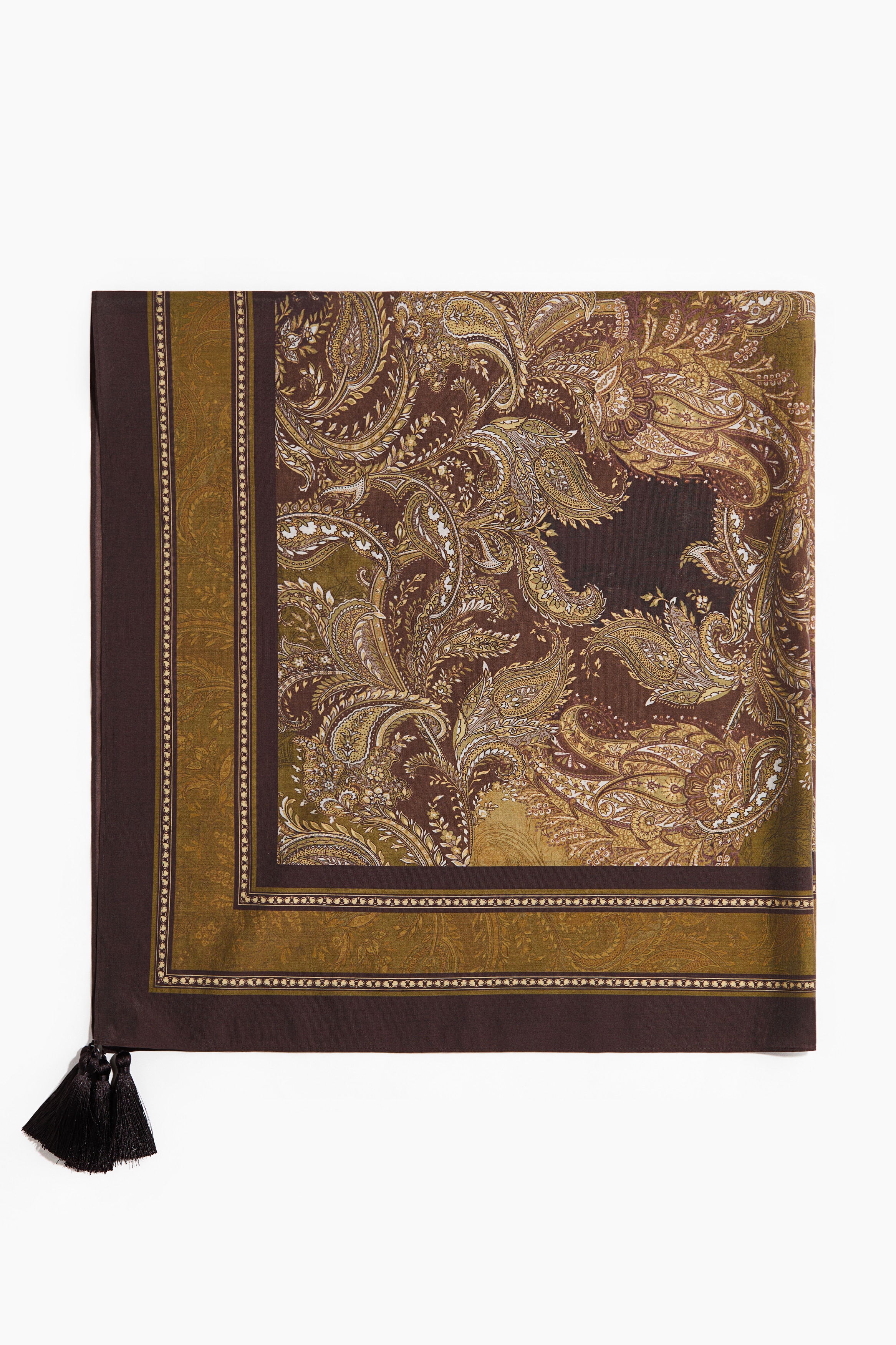 A large square scarf with a brown and olive green paisley print is worn tied around the modelâs waist over a yellow top and black trousers. The scarf features small dark tassels hanging from its corners.