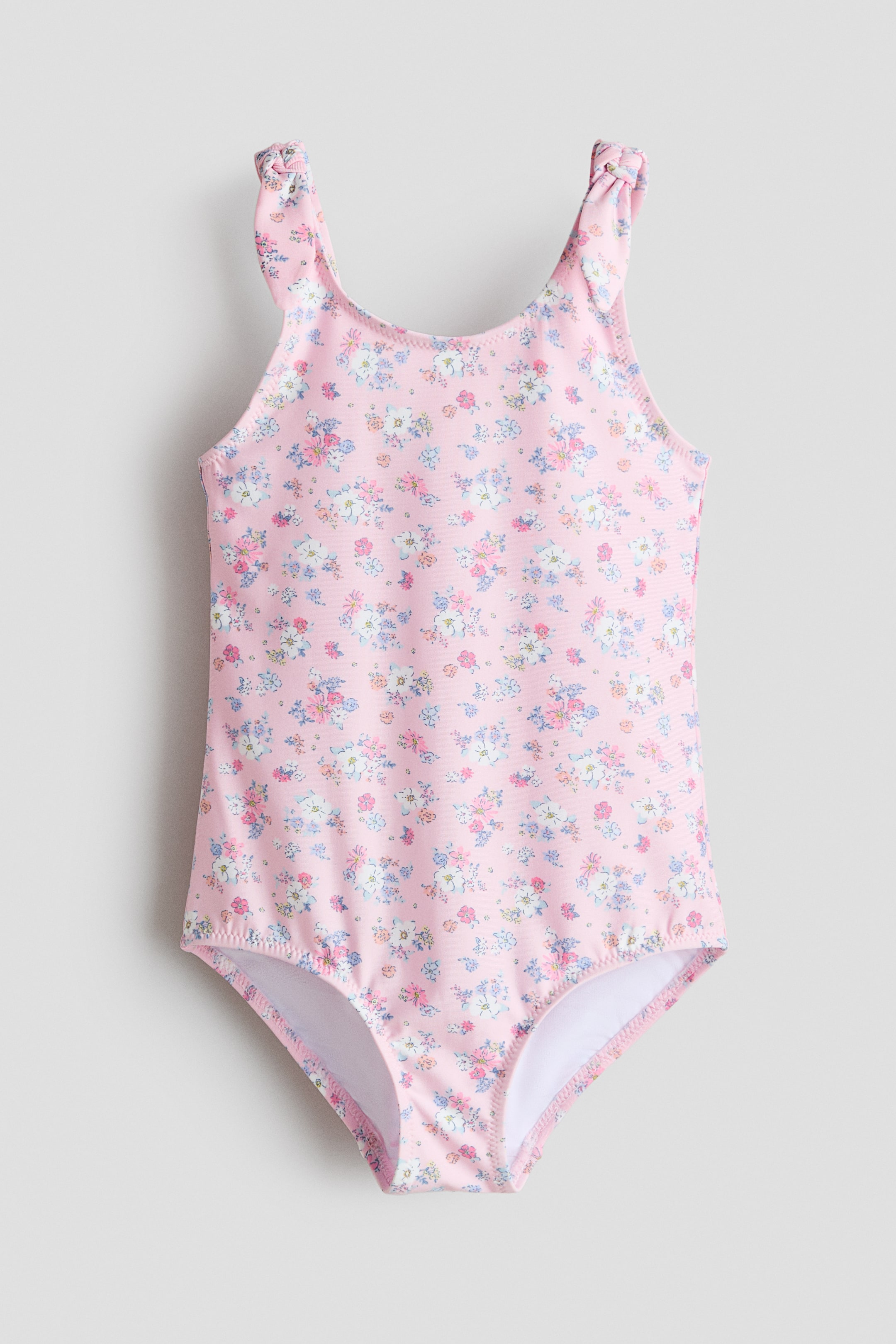 查看大圖: 一件粉色泳裝以正面（FRONT）呈現，表面覆有整件花卉印花。肩帶帶有裝飾性結飾細節，褲腿口處可見白色裡襯。