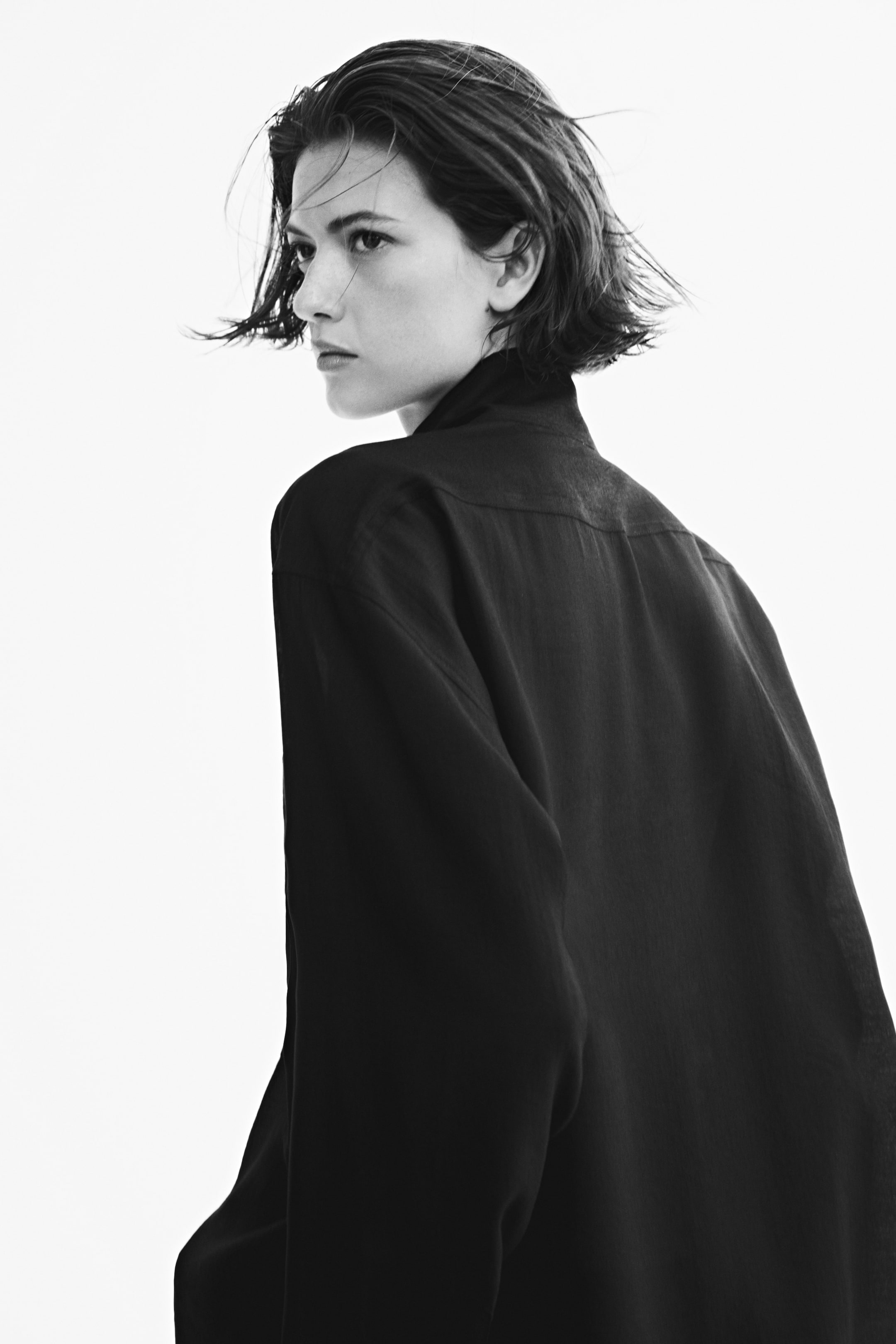 View larger image: An oversized black shirt with a stand collar and a back yoke is being worn by the model. The loose-fitting garment features dropped shoulders and a subtle sheen, shown from a back-angle view as the model looks over their shoulder.