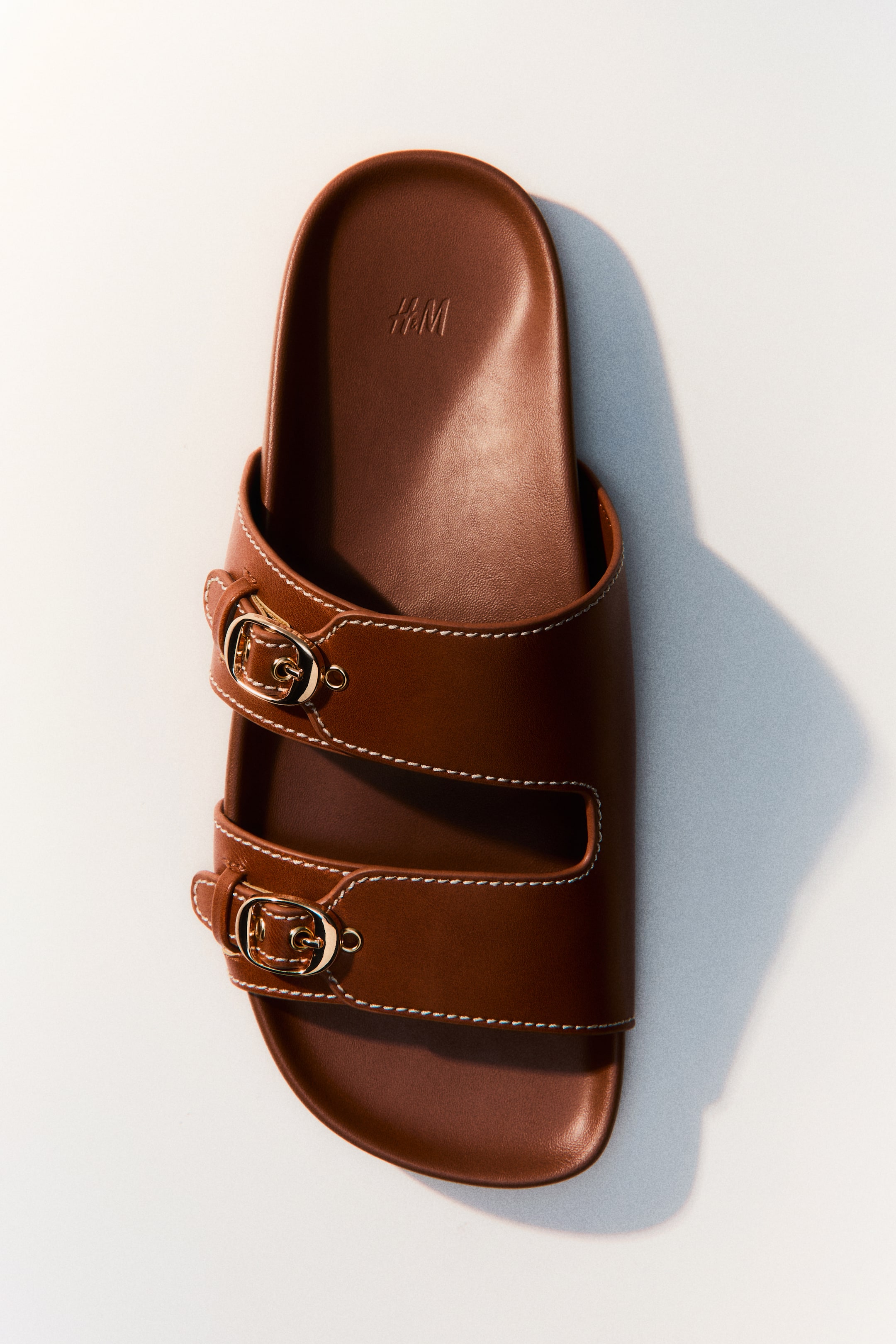 View larger image: The image displays a brown slide sandal from a top-down angle, highlighting two wide straps with gold-toned buckles and white contrast stitching. Its contoured footbed features a debossed H&M logo near the toe.