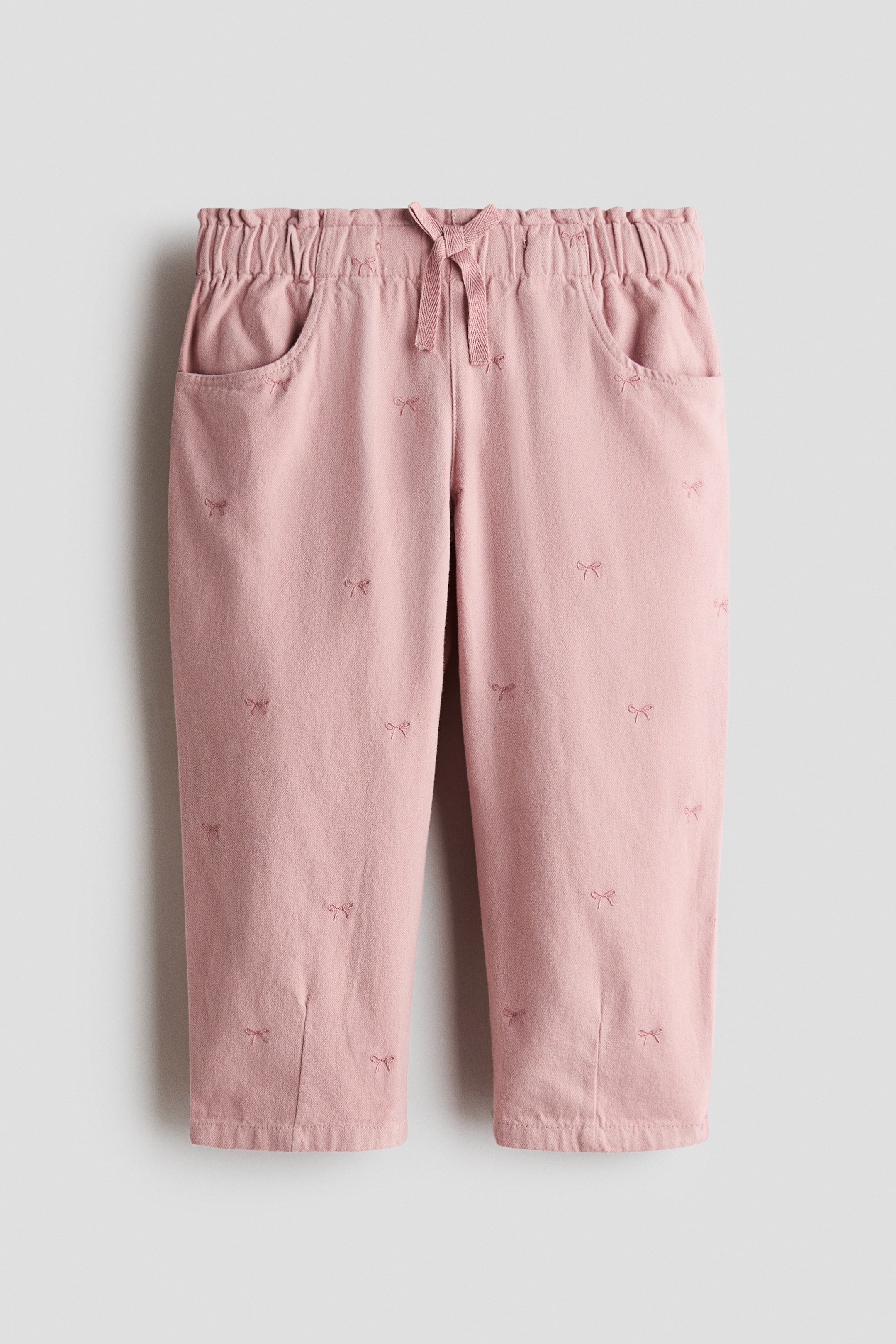 Grotere afbeelding bekijken: Deze lichtroze broek is van voren weergegeven en toont een all-over patroon van kleine geborduurde strikjes. Ze heeft een elastische, gerimpelde tailleband met een centraal trekkoord en gebogen zijzakken.