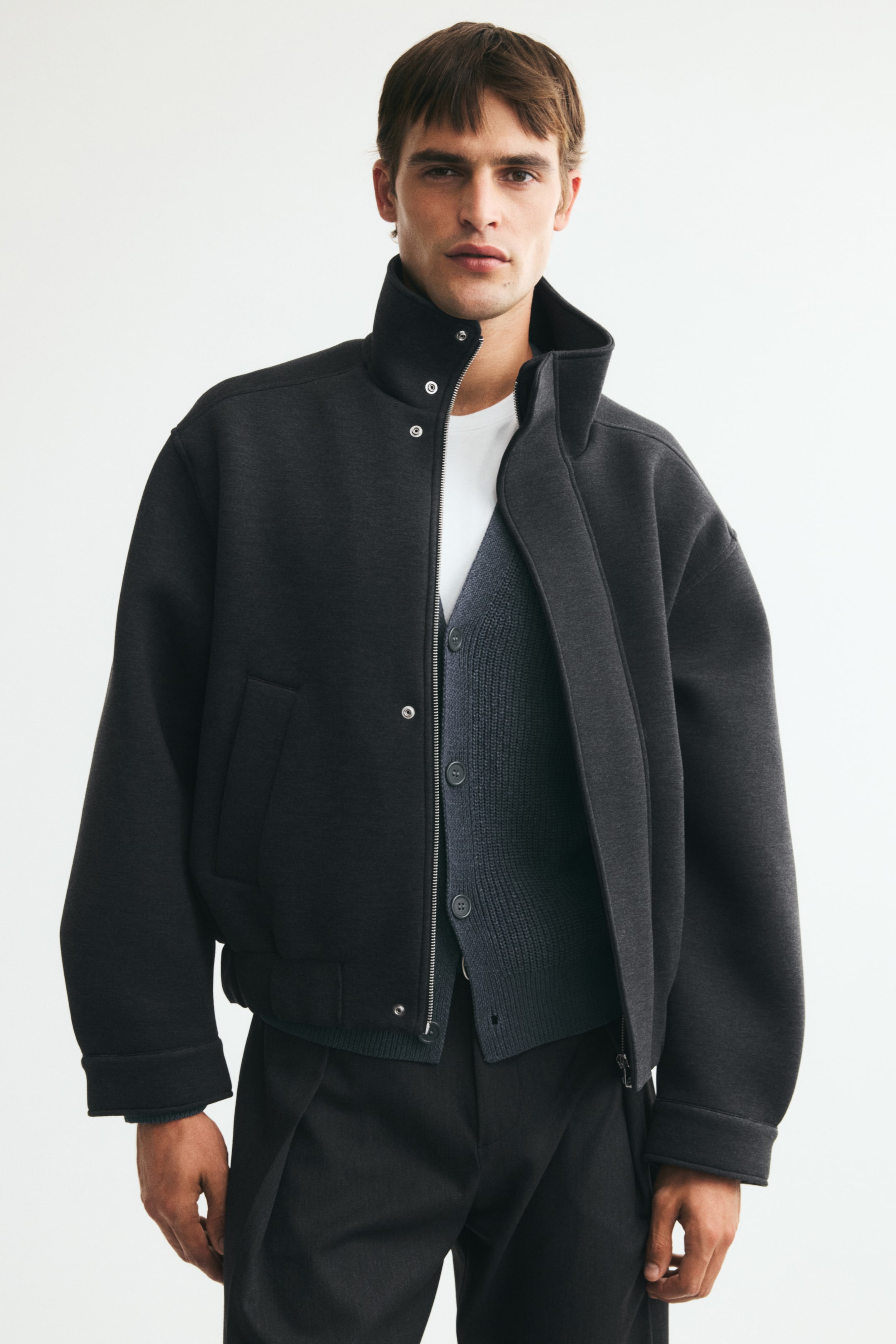View larger image: A dark grey bomber jacket, made from a structured fabric, is being worn by the model. It features a stand-up collar with snap buttons, a partially unzipped front, and side pockets, while layering over a white T-shirt and a dark grey button-up cardigan.