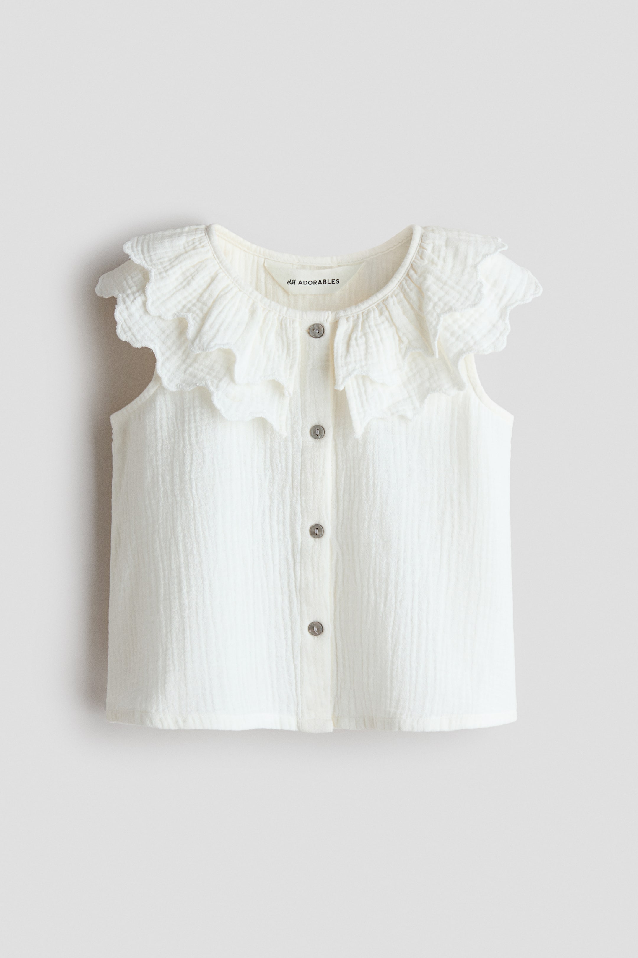 Ver imagen más grande: Una blusa sin mangas H&M Adorables se presenta de frente, con un tejido texturizado en color blanco roto y un escote redondo. Un cuello de doble capa con volantes y bordes festoneados enmarca el escote y se extiende sobre los hombros, con botones que recorren el centro del frente.