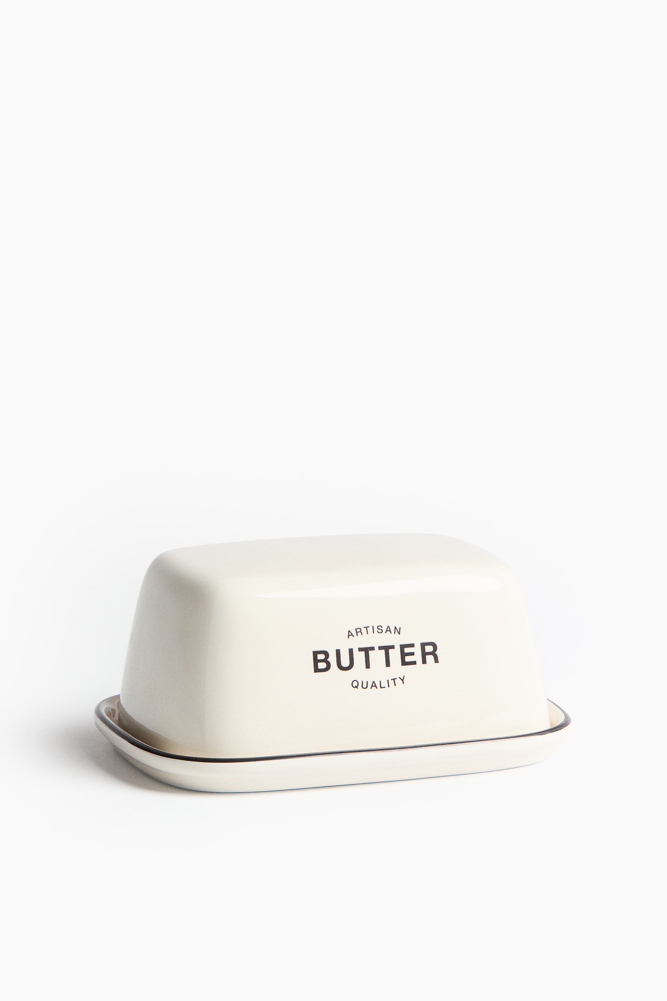 View larger image: The butter dish in a soothing cream hue showcases its front, adorned with a dome-shaped lid inscribed with "ARTISAN BUTTER QUALITY" in bold black lettering, elegantly placed on a rectangular tray. Both the lid and the tray are accentuated with a delicate black trim along their edges, offering a refined touch.