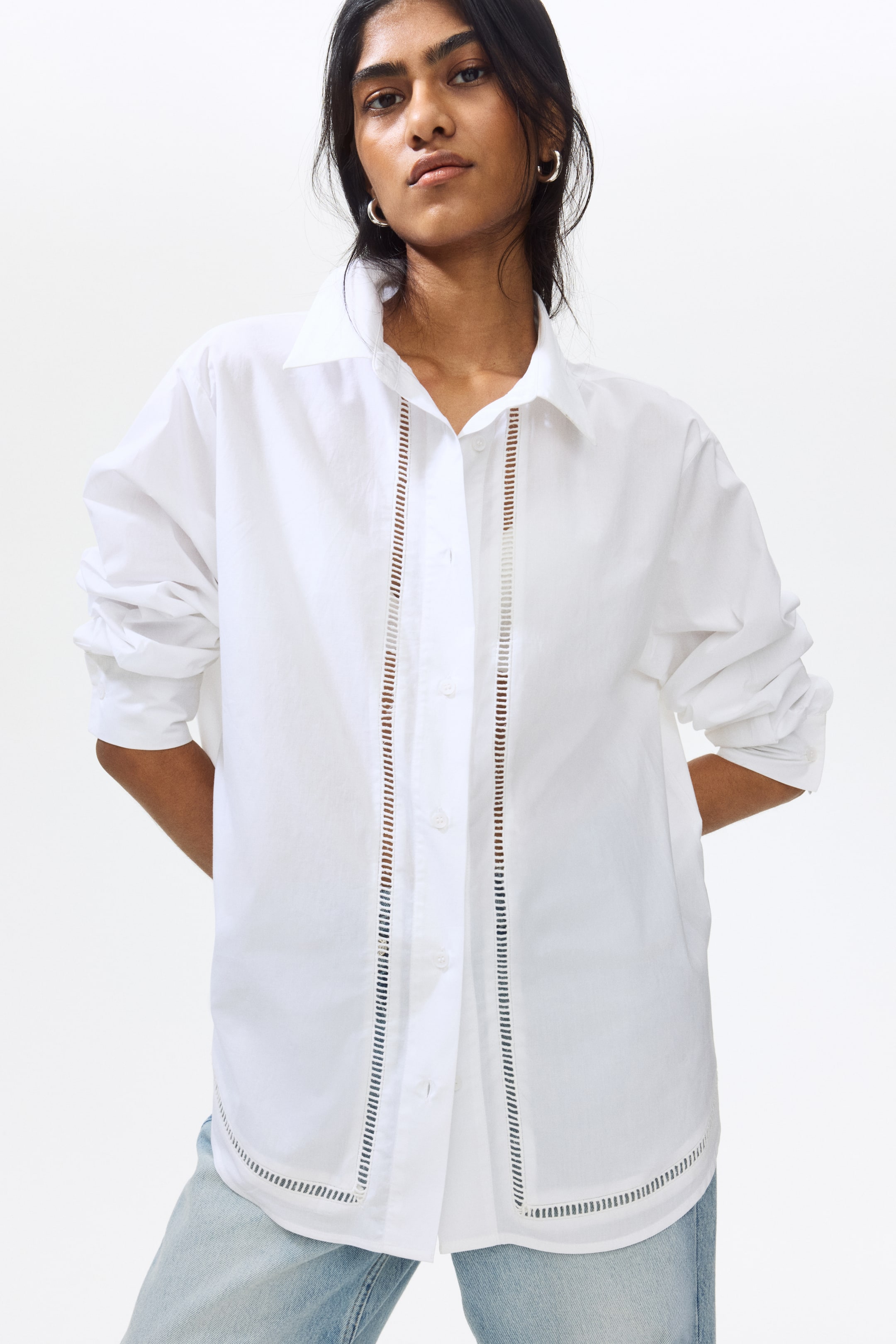 View larger image: The white button-down shirt is showcased from the front, highlighting a pointed collar and vertical ladder-cut detailing on both sides of the front placket. Similar ladder-cut detailing is also visible along the curved hemline as the shirt is paired with light wash jeans, with its long sleeves styled pushed up.