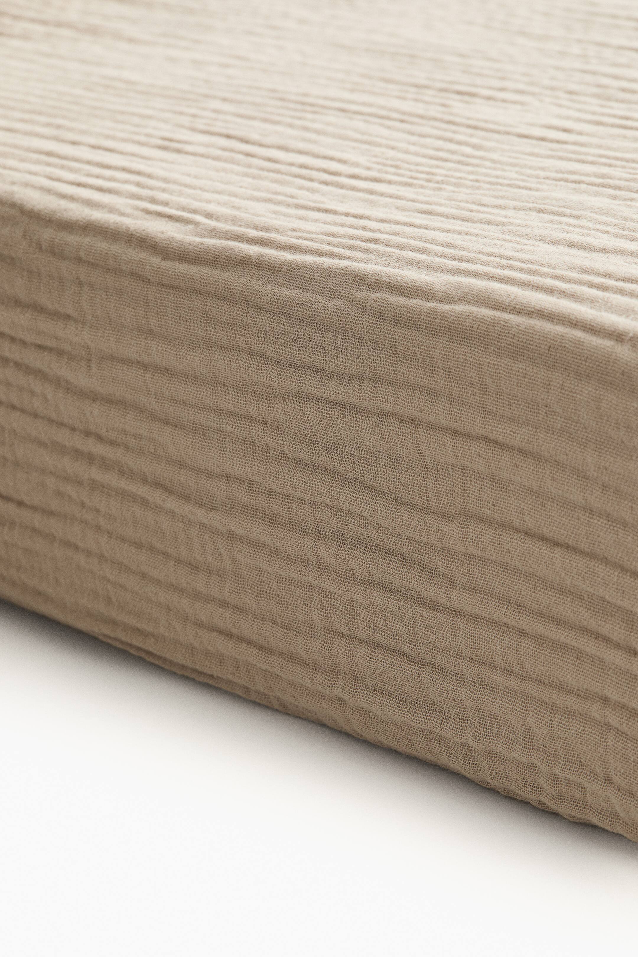 View larger image: A crib fitted sheet in light beige cotton muslin is covering the corner of a mattress, showcasing its snug fit and soft, crinkled texture.