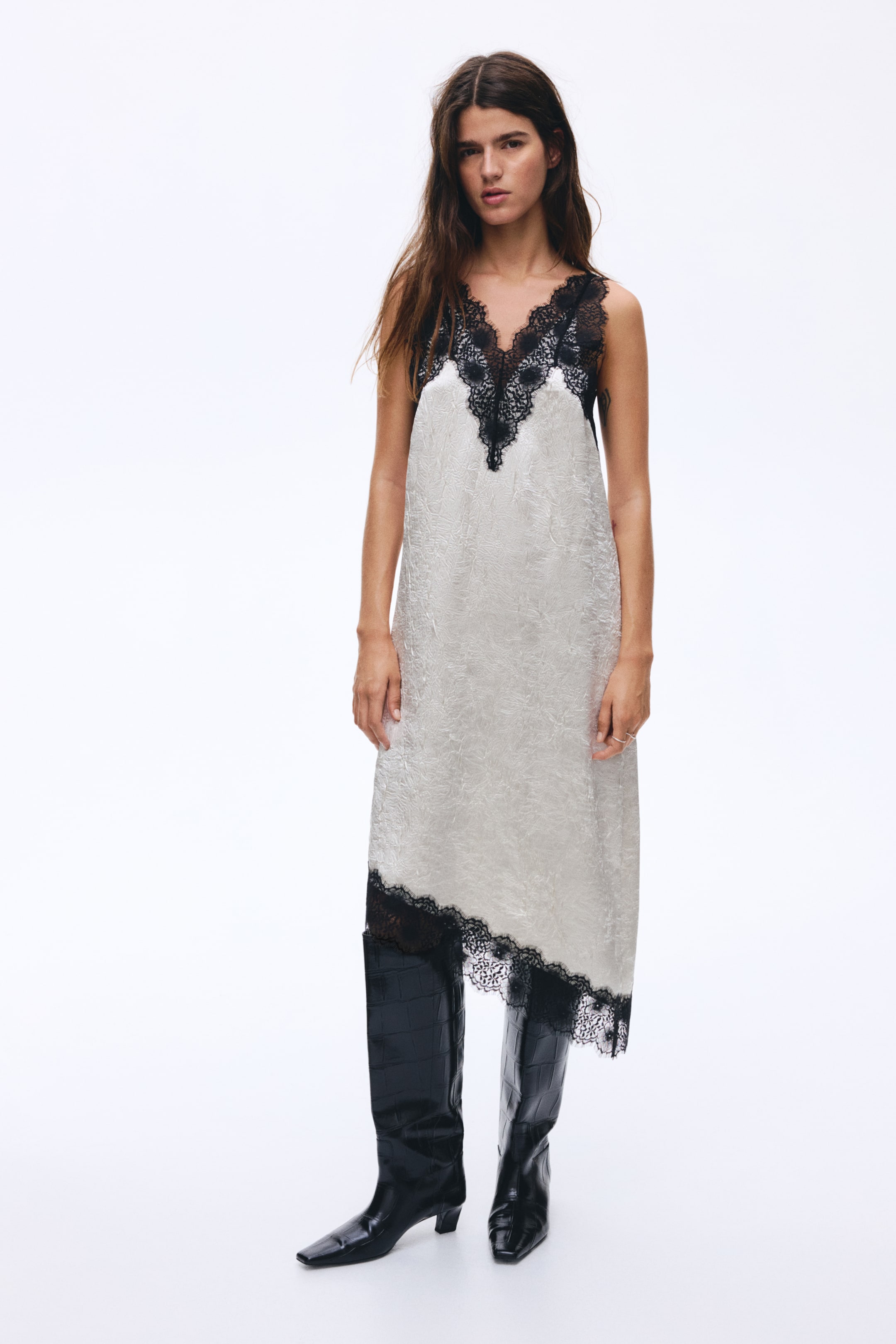 Zobraziť väčší obrázok: Svetlá slip šaty sú prezentované z predného pohľadu, vyrobené z reliéfnej látky so subtílnym vzorom. Sú navrhnuté s čiernou čipkou pozdĺž výstrihu do V a asymetrickým lemom, a voľne padajú cez čierne čižmy po kolená.