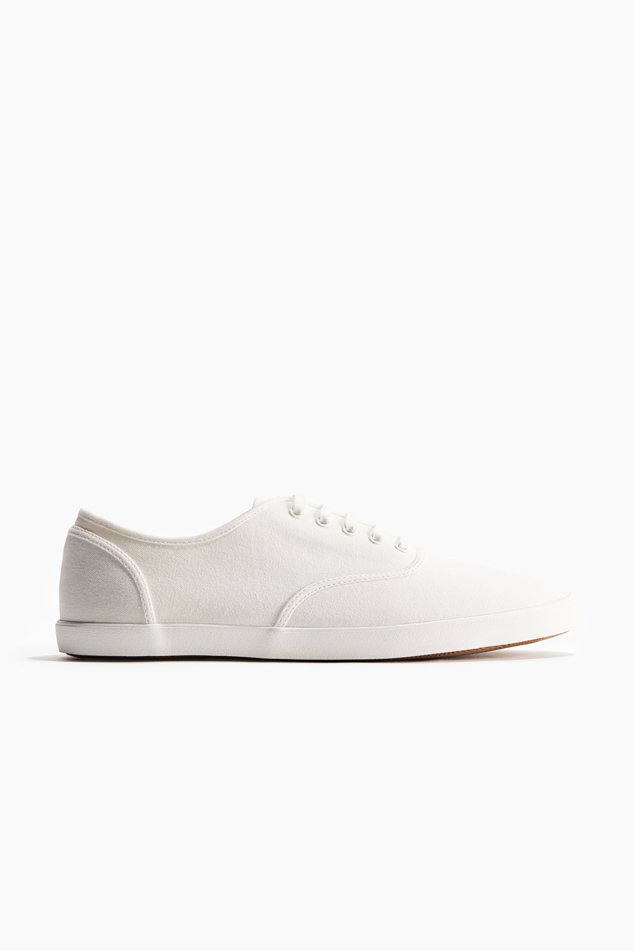 View larger image: A white canvas low-top sneaker features white laces threaded through five pairs of metal eyelets. It showcases a white rubber foxing tape around its perimeter, with a light brown outsole visible at the toe.