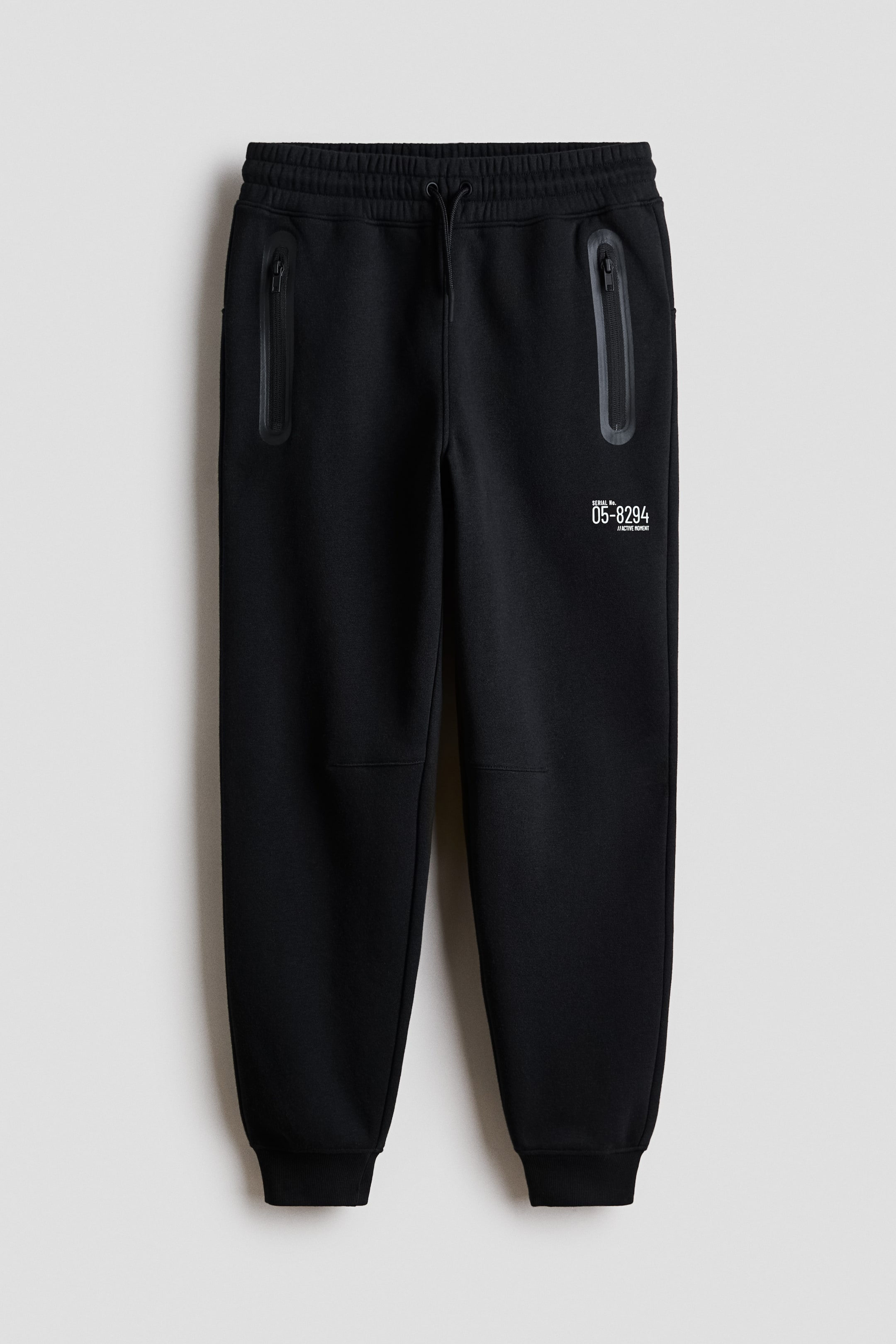View larger image: Black sweatpants feature an elasticated drawstring waistband, two zippered side pockets with sealed trims, and ribbed ankle cuffs. A white serial number graphic is visible on the upper right leg.