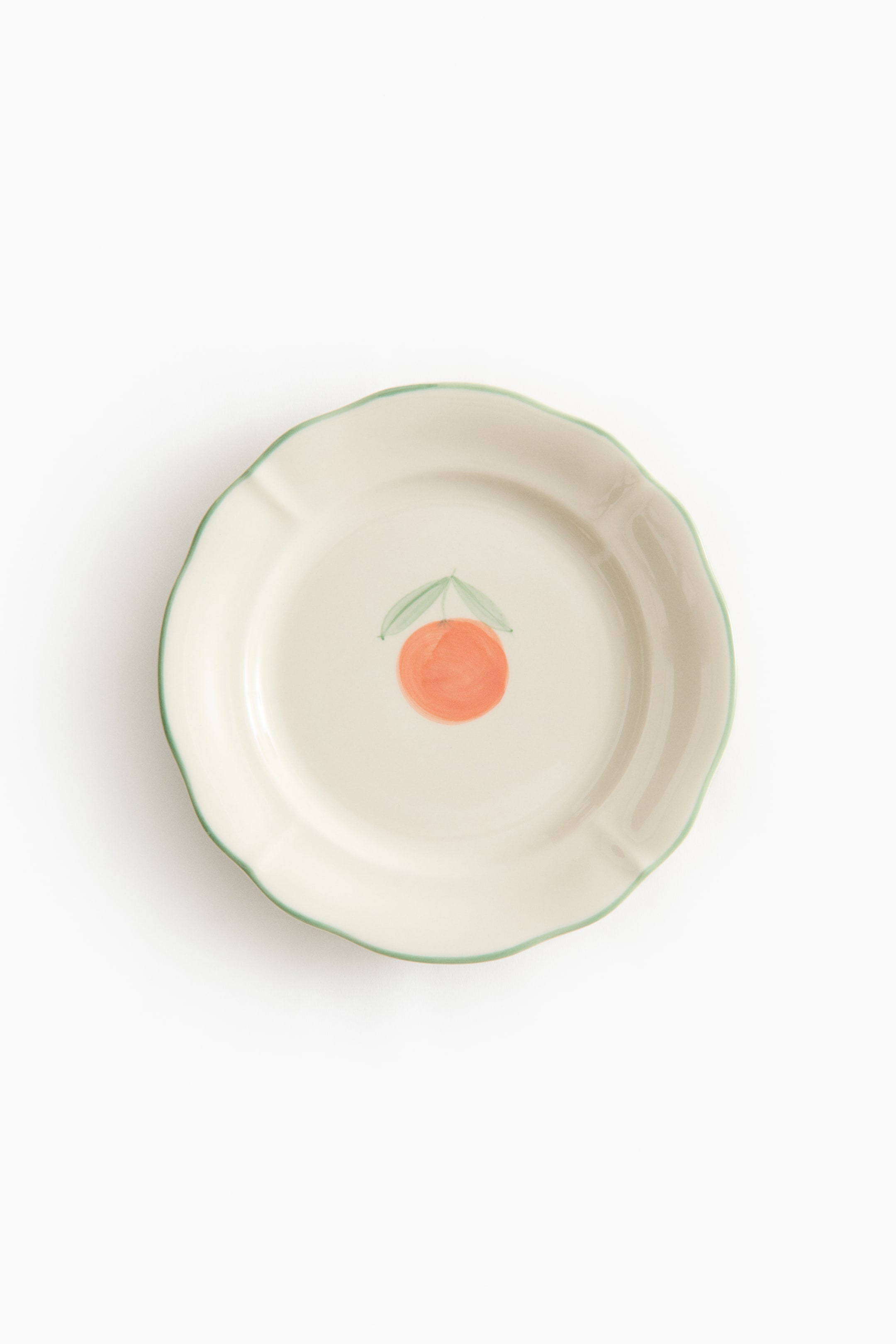 View larger image: A stoneware starter plate faces forward, showing an off-white surface with a scalloped, hand-painted green rim. A hand-painted orange fruit motif with two green leaves is placed centrally on the plate.