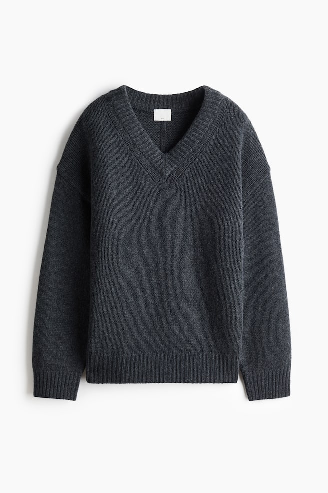 Un pull en laine tricotée gris foncé est présenté de face, avec une coupe oversize, un col en V côtelé et des épaules tombantes. Le modèle met en valeur des manches longues avec des poignets côtelés et un ourlet côtelé assorti. - 1