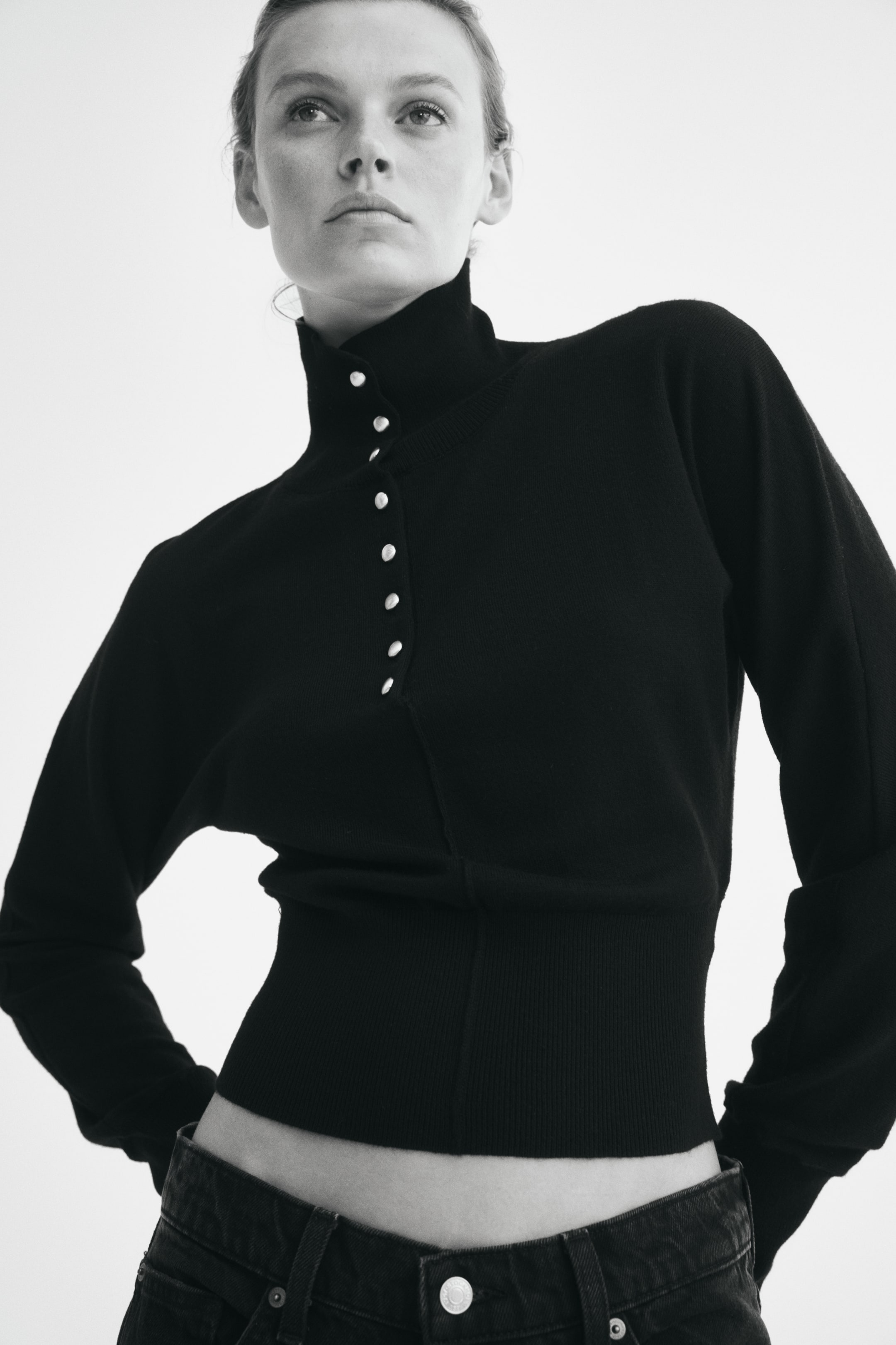 Agrandir l'image: Un pull court noir en maille fine, doté d’un col montant avec une patte verticale ornée de boutons perlés, est porté par le mannequin. Ce pull à manches longues présente un large bord-côte et est associé à un jean de couleur foncée.