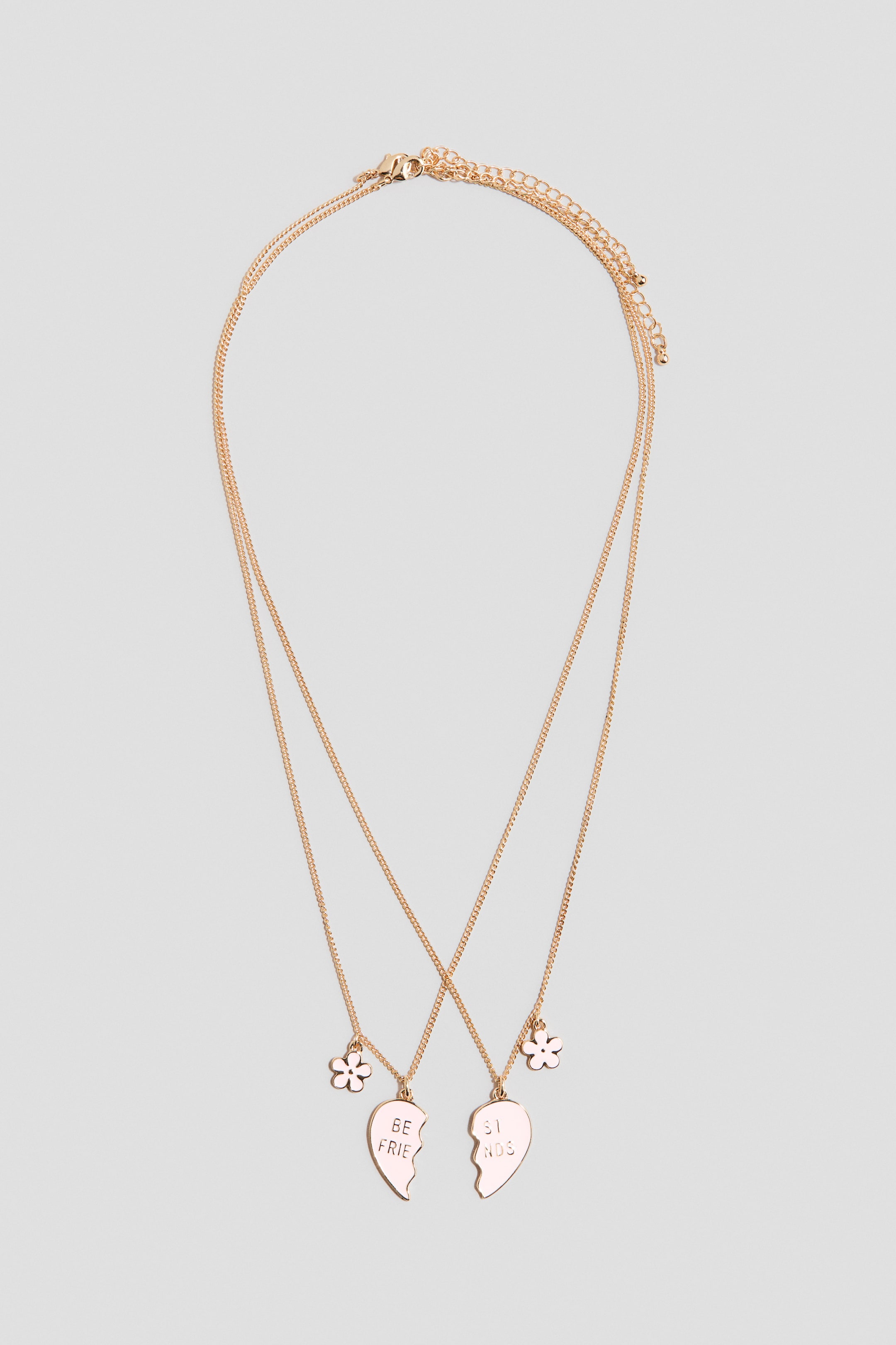 Zobacz większy obraz: Dwa naszyjniki z metalowym łańcuszkiem w kolorze różowego złota prezentują pasujące zawieszki w kształcie serca, z których jedna ma napis „BE FRIE”, a druga „ST NDS”, w towarzystwie małych kwiatowych zawieszek. Każdy naszyjnik posiada regulowany łańcuszek z zapięciem na karabińczyk.