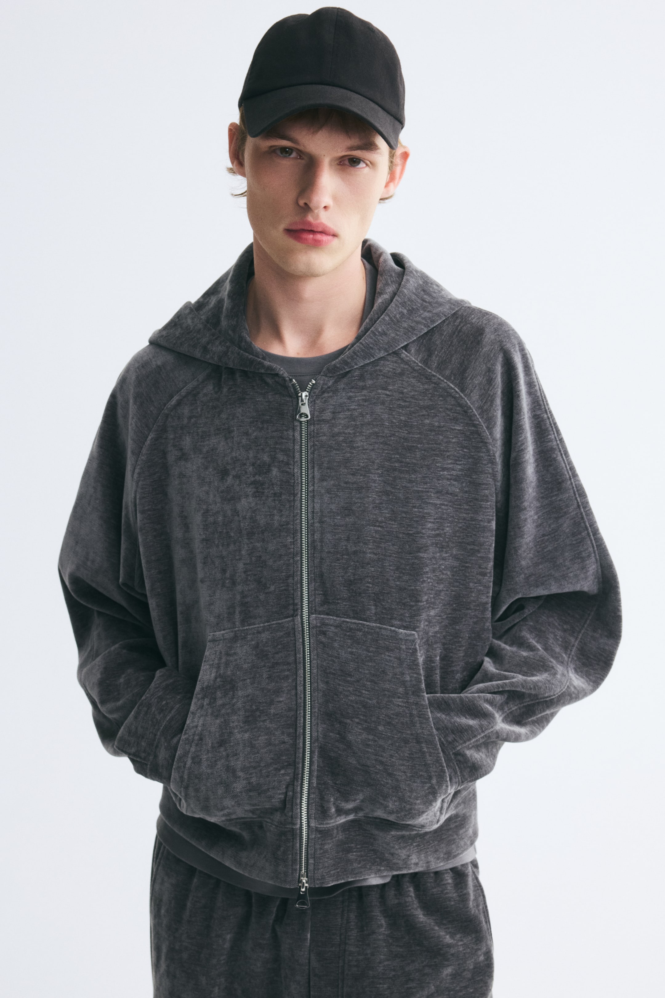 View larger image: A relaxed-fit hooded jacket in dark gray velour, featuring a front two-way zipper and front pockets, is worn by the model with hands in the pockets. The jacket is styled with matching velour trousers, a gray T-shirt, and a black cap.