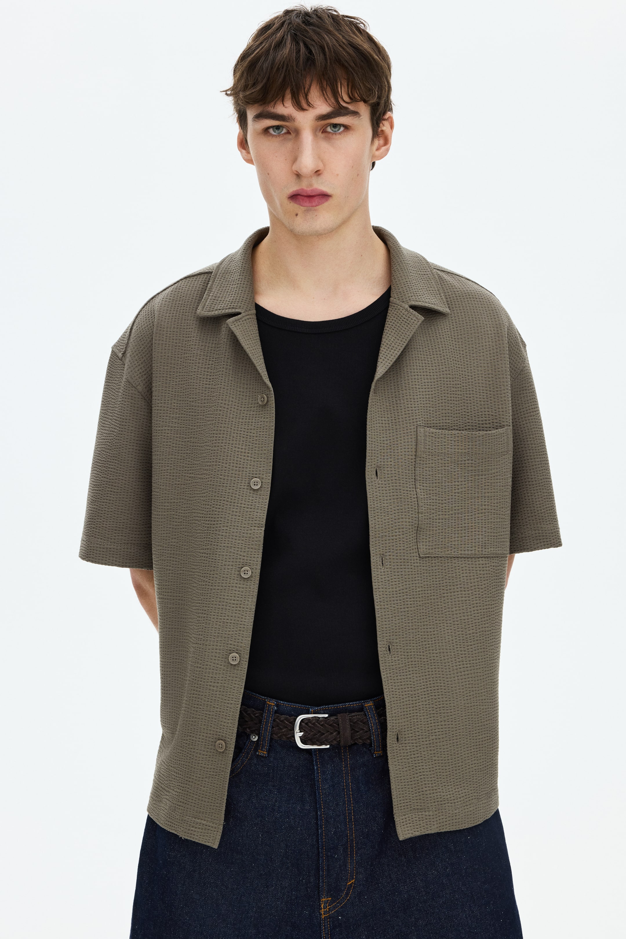 Ver imagen más grande: Una camisa de manga corta en una mezcla de algodón texturizado está siendo llevada por el modelo. La camisa de corte relajado presenta un cuello resort, una parte frontal francesa con botones visibles, un bolsillo en el pecho y un dobladillo recto, y se lleva abierta sobre una camiseta interior negra.