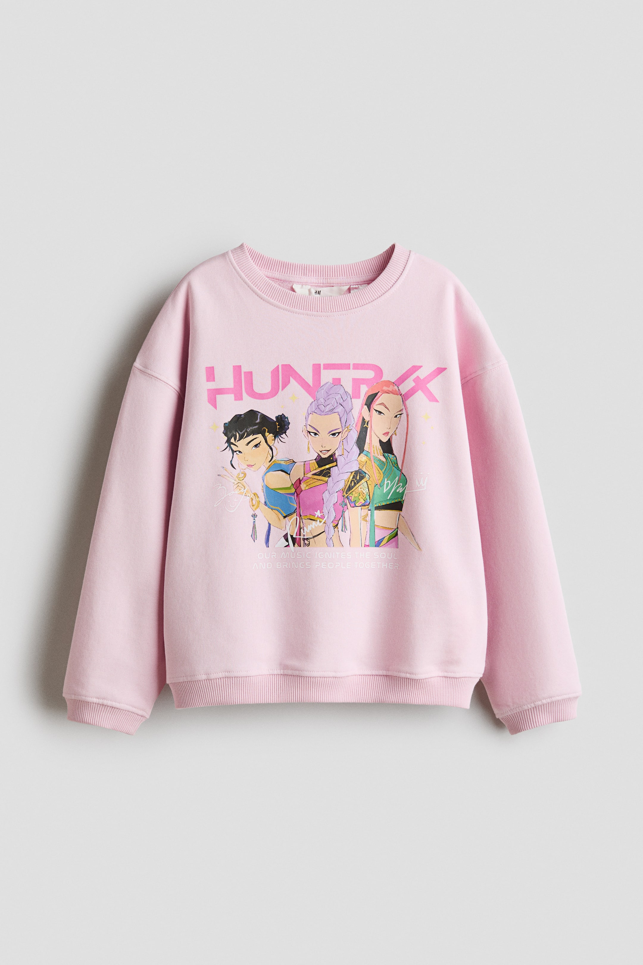 Größeres Bild anzeigen: Ein rosa Sweatshirt wird von vorne präsentiert und zeigt ein farbenfrohes Motiv von drei animierten weiblichen Charakteren mit dem Text "HUNTRXX" darüber und "OUR MUSIC IGNITES THE SOUL AND BRINGS PEOPLE TOGETHER" darunter. Es ist mit einem runden, gerippten Ausschnitt, überschnittenen Schultern, langen Ärmeln sowie gerippten Bündchen und Saum gestaltet.