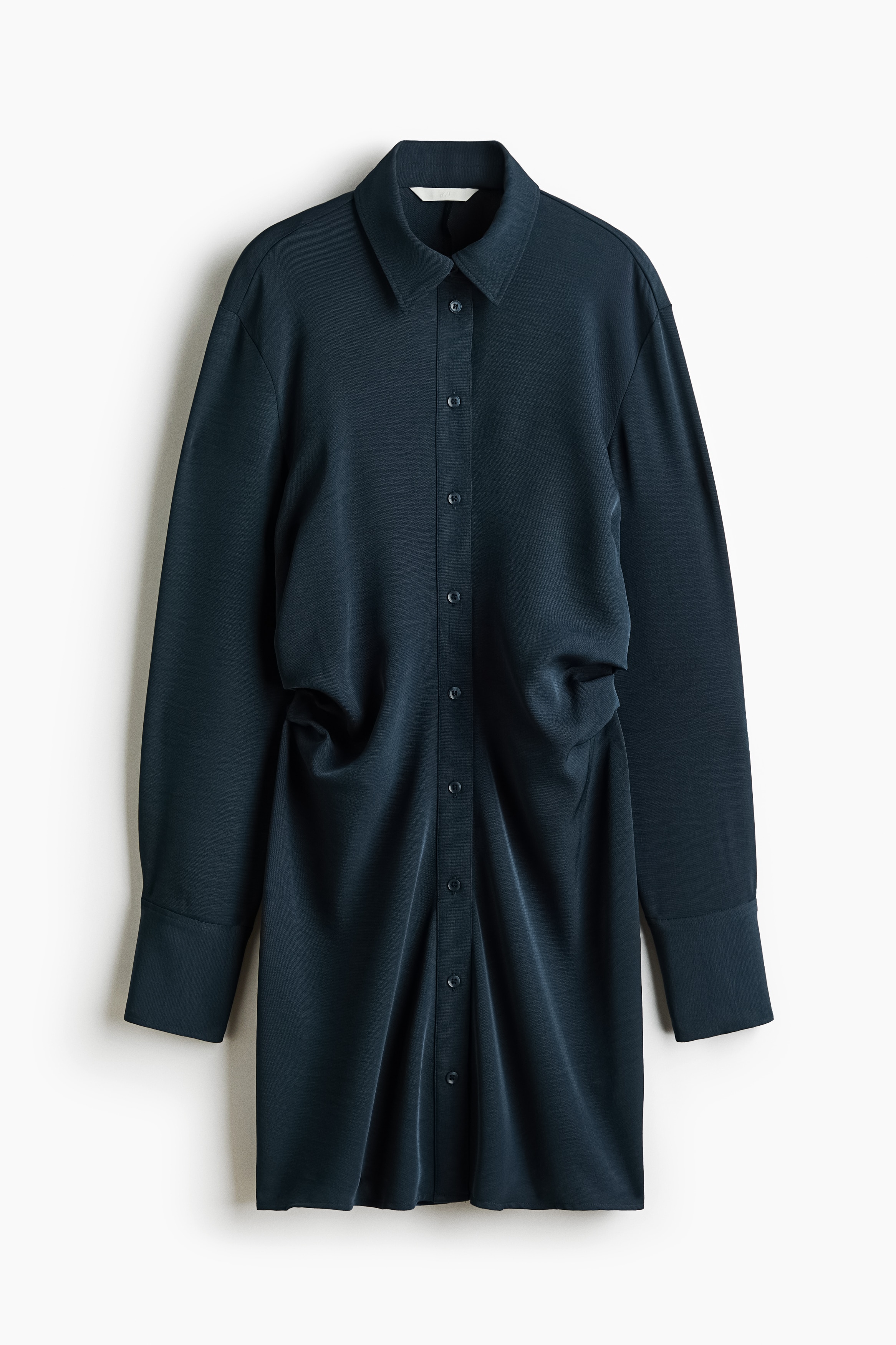 La modelo lleva un vestido camisero ajustado de color azul oscuro, con cuello, cierre de botones al frente y detalle fruncido en la cintura. Posan con un brazo levantado hacia la parte posterior del cuello, resaltando las mangas largas y la silueta drapeada.