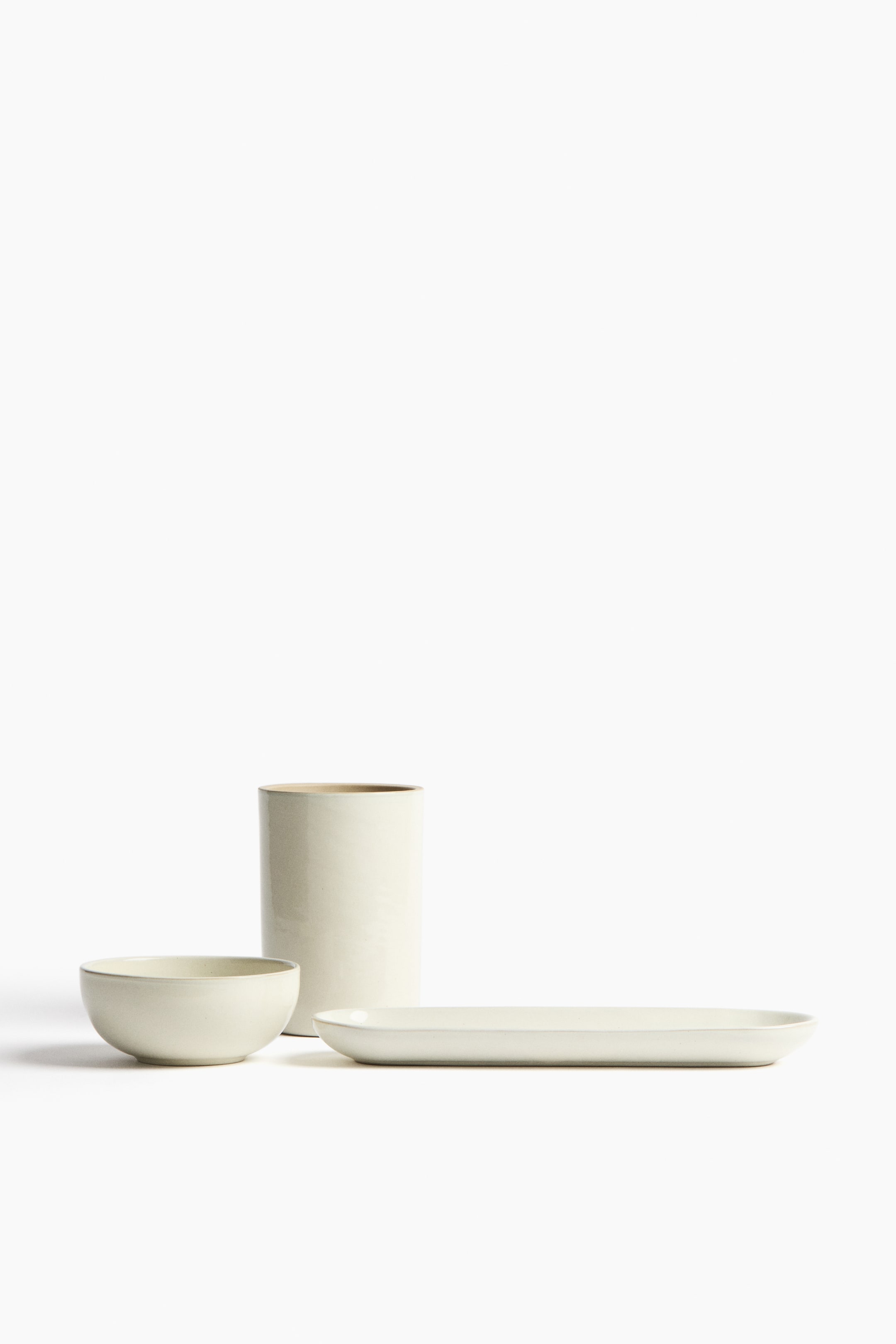 Ver imagen más grande: Una colección de vajilla de cerámica en color crema claro se presenta, con una taza cilíndrica alta, un cuenco redondo y un plato ovalado largo. Cada pieza muestra un sutil acabado mate y está dispuesta de frente.