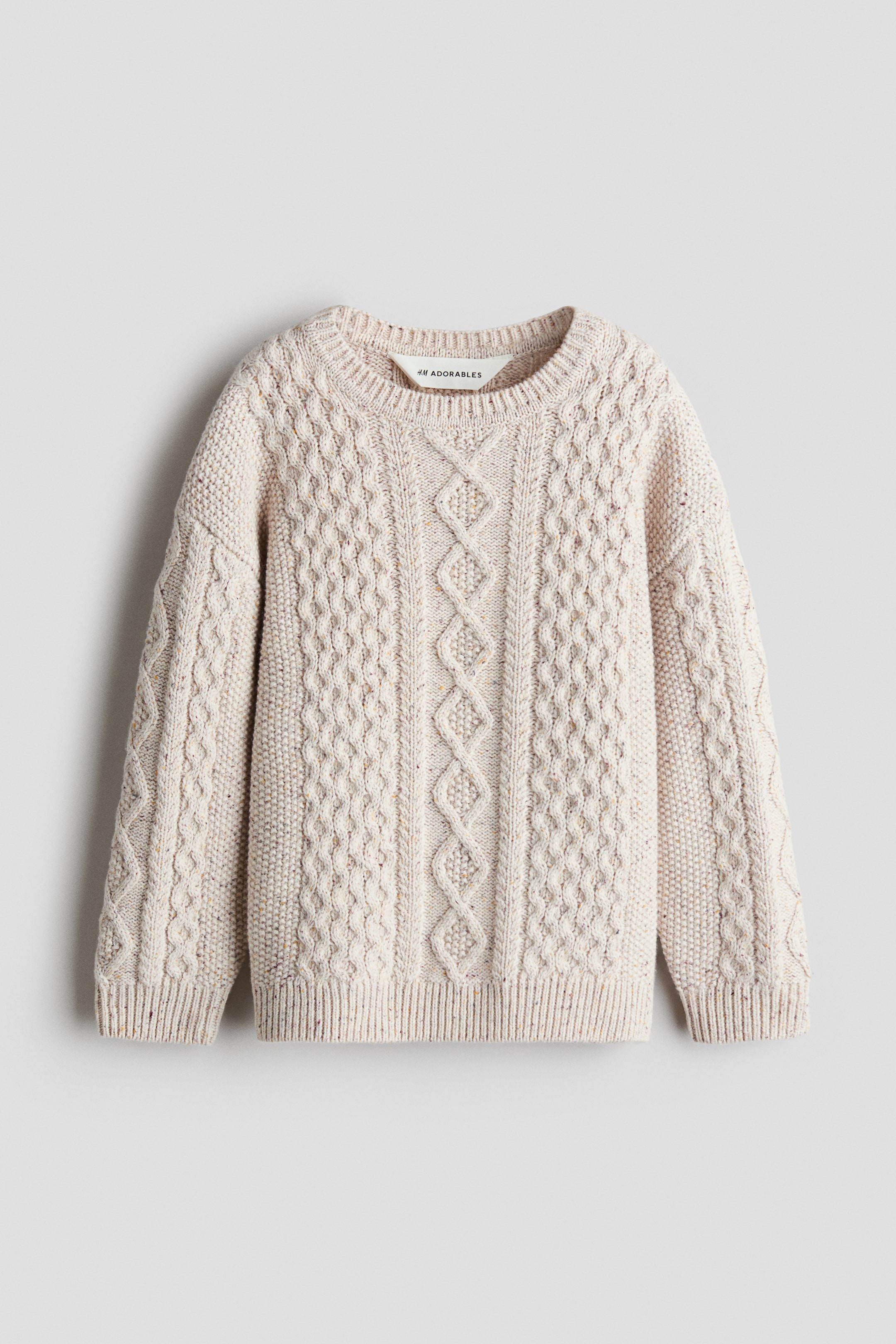 Visa större bild: En kabelstickad tröja från H&M Adorables visas framifrån och framhäver en krämfärgad, melerad ullstickning med intrikata kabelmönster. Den lediga tröjan har en rund ribbad halsringning, nedhasade axlar samt ribbade muddar och fåll.
