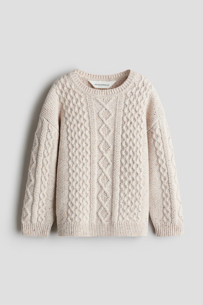 Un pull en maille torsadée H&M Adorables est présenté de face, mettant en valeur une maille de laine crème mouchetée aux motifs torsadés raffinés. Ce pull à la coupe ample présente un col rond côtelé, des épaules tombantes, ainsi que des poignets et un ourlet côtelés. - 1