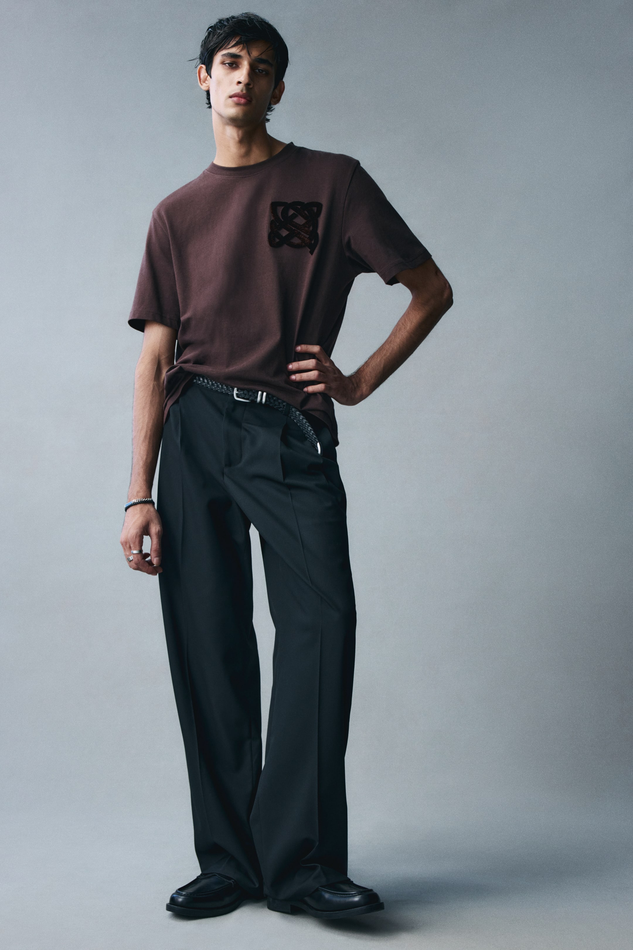 View larger image: A brown cotton jersey T-shirt with a round neckline, short sleeves, and a black graphic print on the left chest is worn by the model. The regular-fit tee is styled with a partial tuck into wide-leg black trousers, finished with a braided belt.