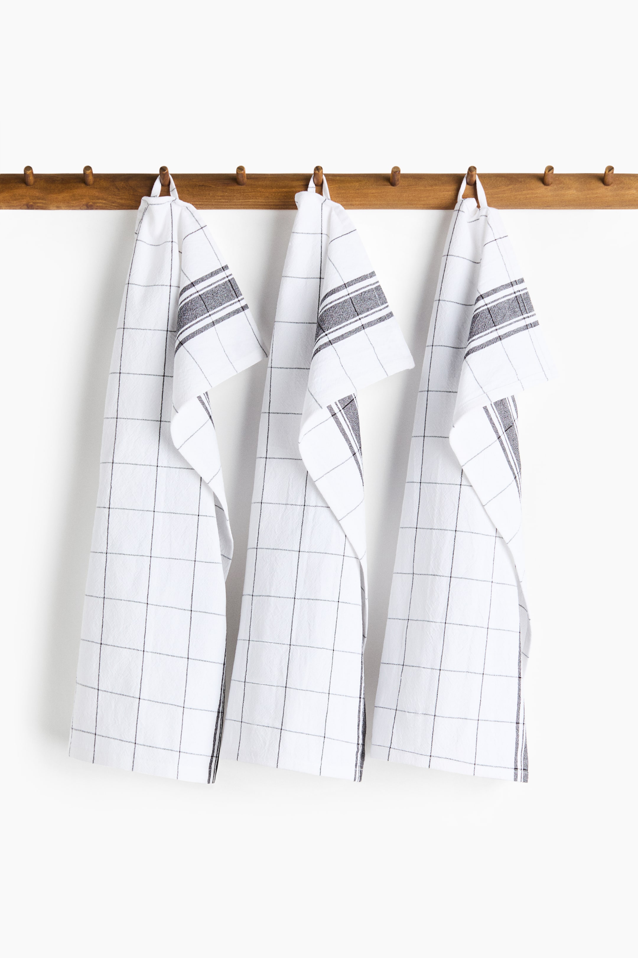 View larger image: A set of three cotton tea towels is hanging, showcasing a white fabric with a black grid pattern. Each towel also features a dark grey striped band at one end and a hanger on one short side.
