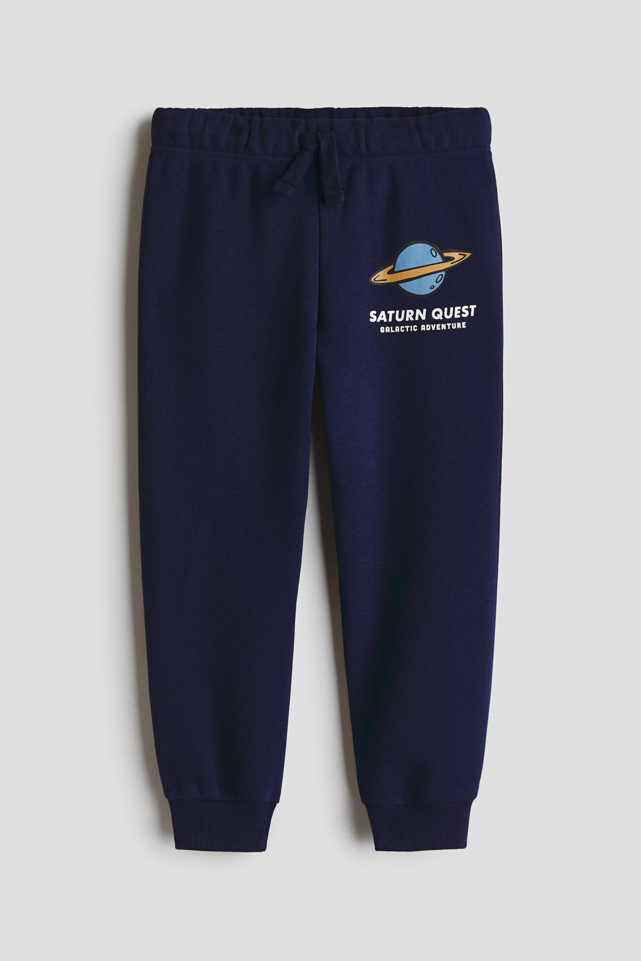 View larger image: Front-facing navy blue joggers featuring an elasticated drawstring waist and ribbed cuffs. A graphic print of Saturn with rings and the text 'SATURN QUEST GALACTIC ADVENTURE' appears on the left leg.