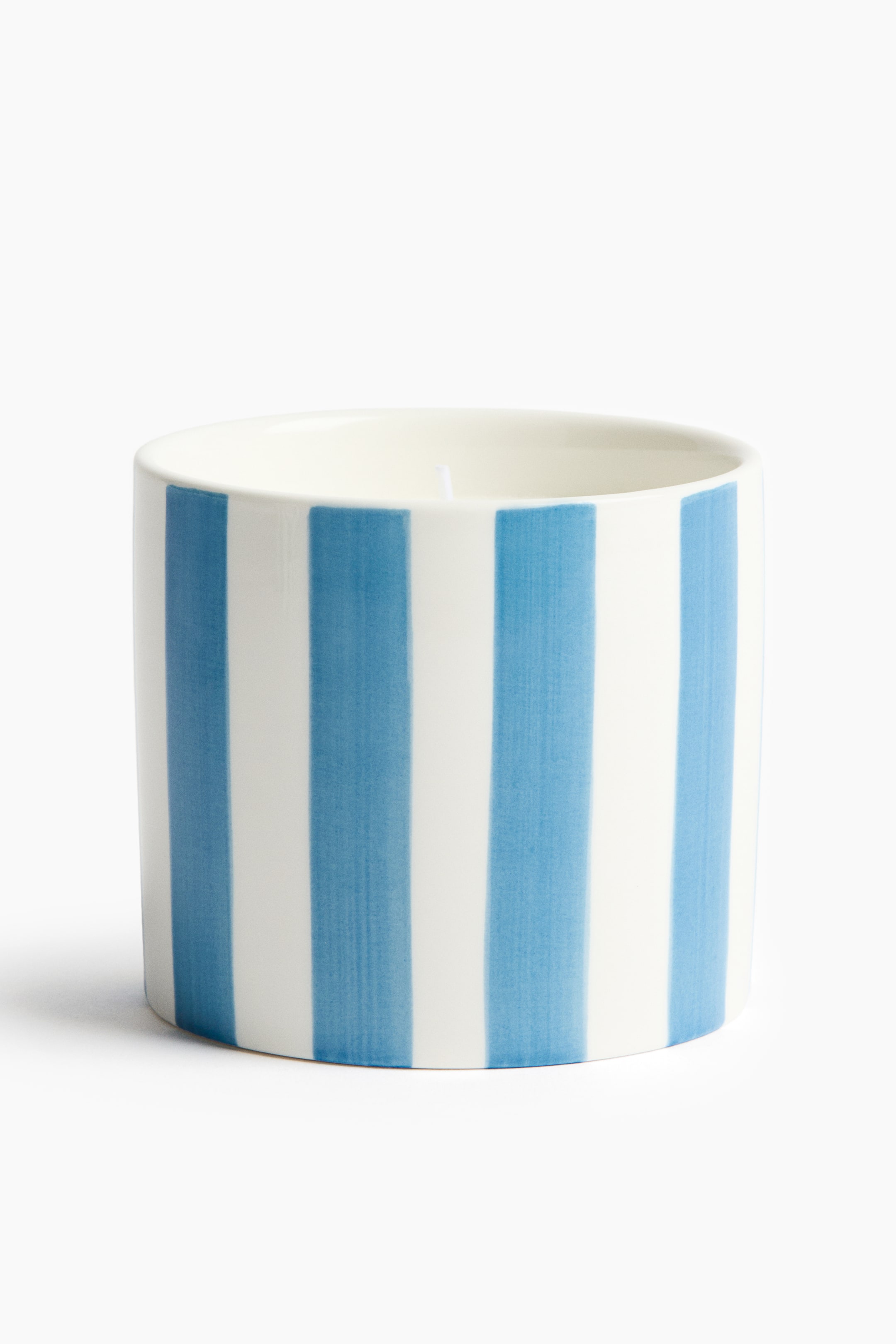 Visa större bild: Ett ljus är inneslutet i en cylindrisk vit keramikkärl, som har handmålade vertikala blå ränder runt dess utsida. Vitt vax med en synlig veke fyller kärlet.