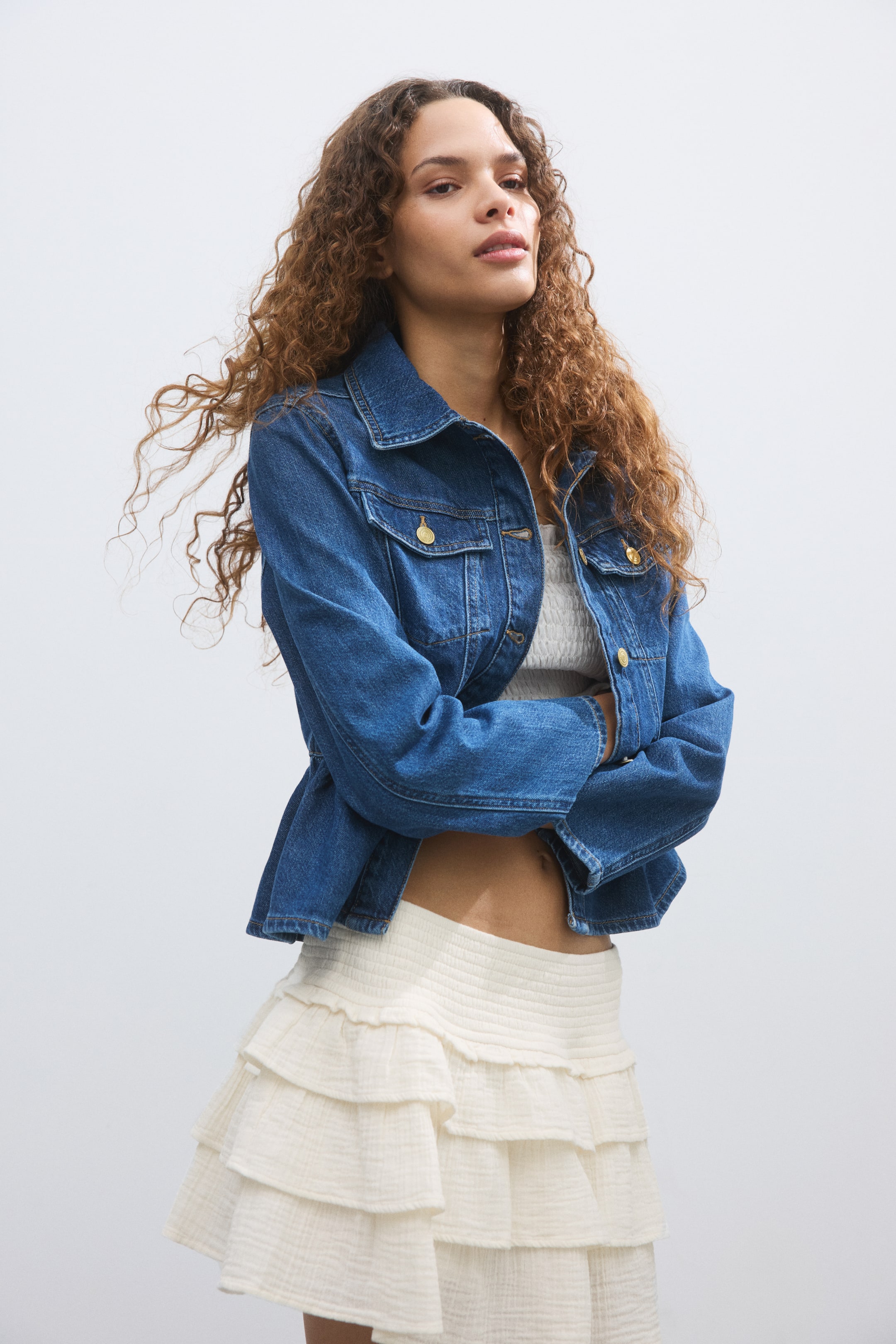 Agrandir l'image: Une veste en jean bleu moyen présente un col classique, deux poches à rabat sur la poitrine avec des fermetures à boutons dorés, et une patte de boutonnage avant avec des boutons assortis. La veste est portée ouverte sur un haut blanc, mettant en valeur sa coupe légèrement raccourcie et ses manches longues.