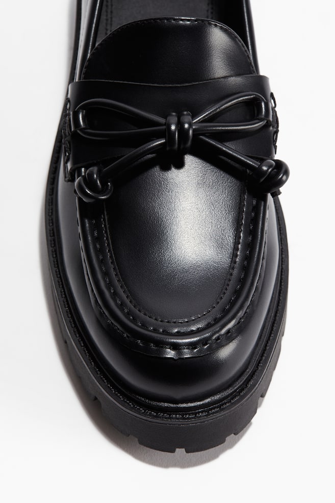 Un mocassin noir présente un bout arrondi avec des coutures mocassin, une semelle épaisse à crampons et un nœud décoratif noué sur sa bride de cou-de-pied. - 1
