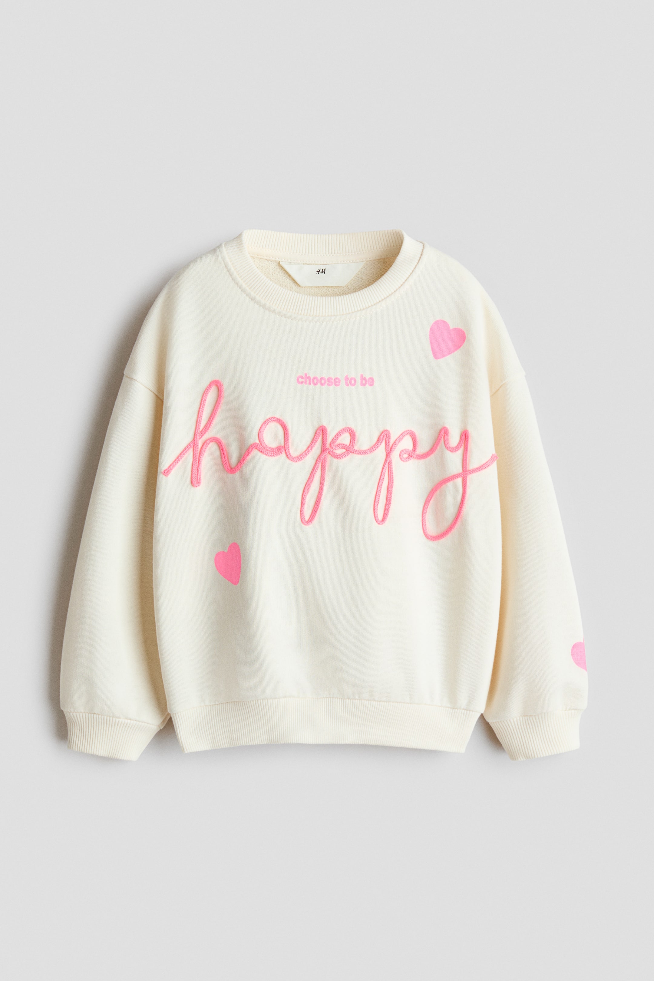 View larger image: A cream-coloured sweatshirt is shown from the front, featuring a ribbed crew neck, dropped shoulders, and ribbed cuffs and hem. The front features the word "happy" in raised pink cursive embroidery, with the phrase "choose to be" printed in pink above it and small pink hearts printed on the front and sleeve.