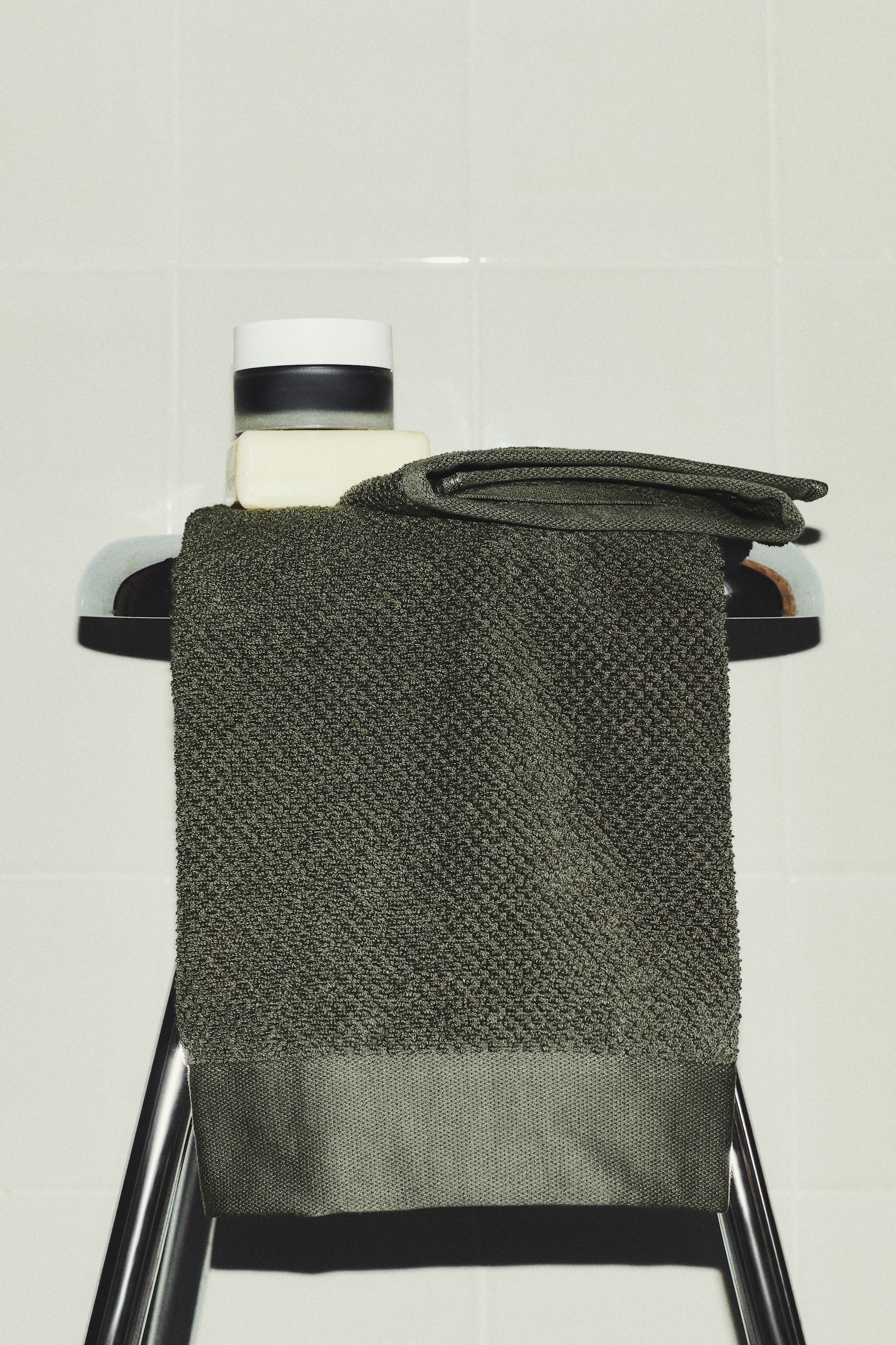 Agrandir l'image: Une serviette vert olive est drapée sur une barre à serviette chromée, mettant en valeur son corps principal texturé et une bordure inférieure plus lisse. Un savon et un pot cosmétique reposent sur sa section supérieure pliée.