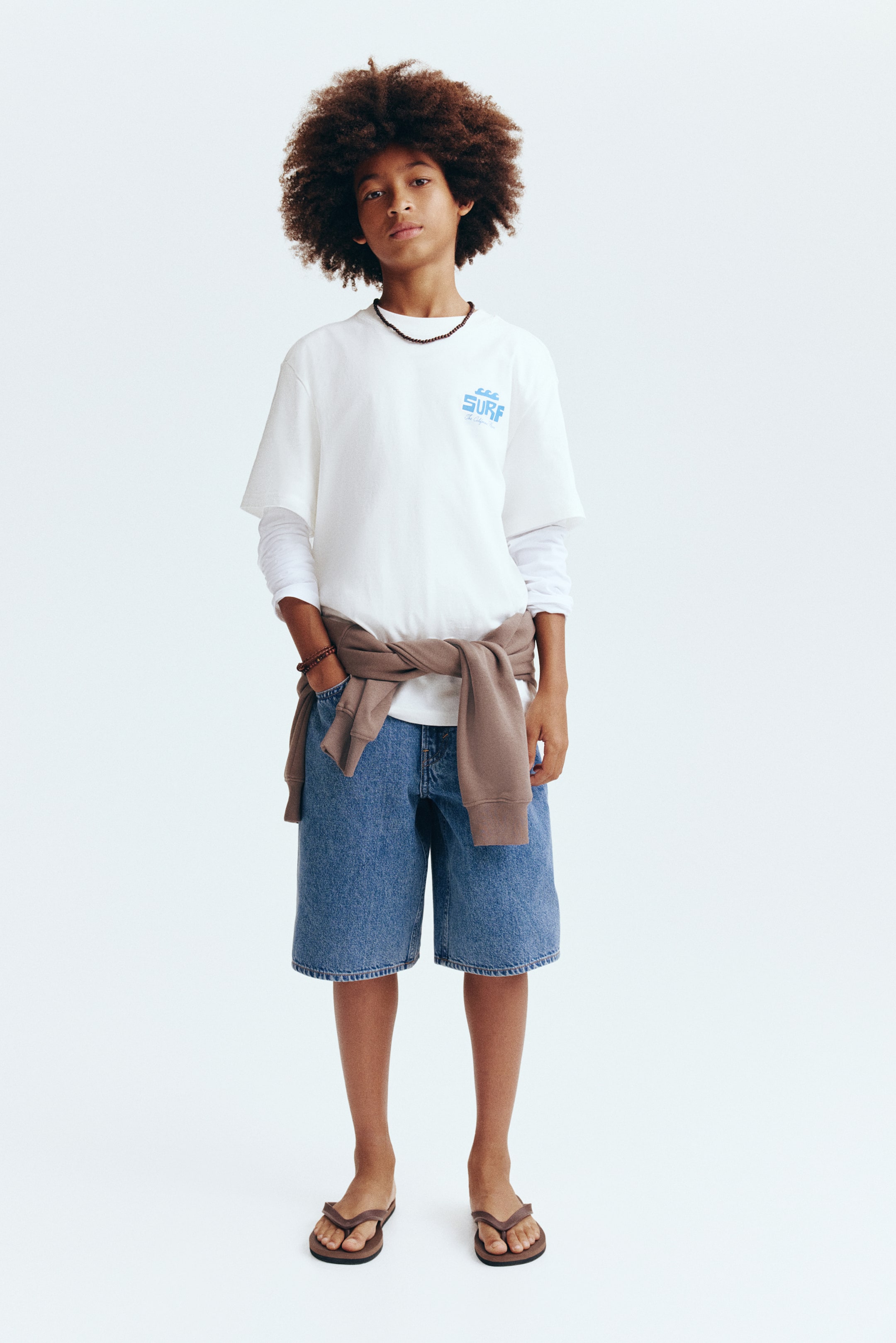 拡大画像を表示: 白いクルーネックの半袖Tシャツが正面を向いており、左胸に波のアイコンと一緒に青い「SURF」グラフィックが描かれています。白い長袖シャツの上に重ね着されており、袖が下から伸びています。