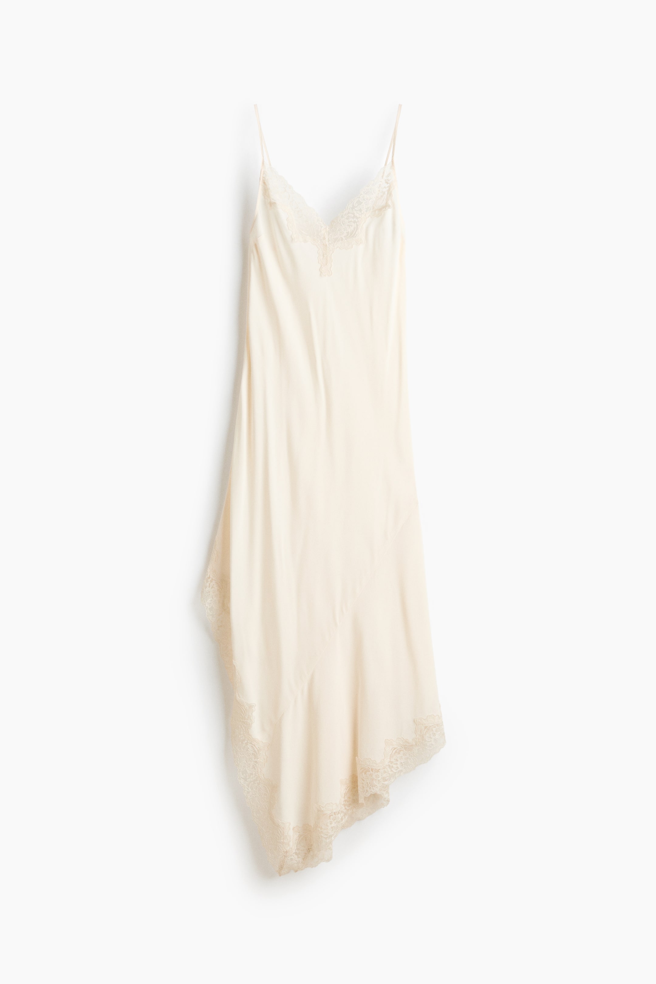 Größeres Bild ansehen: Ein cremefarbenes Slip-Kleid präsentiert seine Vorderansicht, mit einem V-Ausschnitt, der mit Spitze besetzt ist, und dünnen Spaghettiträgern. Der fließende Stoff formt einen asymmetrischen Saum, der ebenfalls mit Spitze verziert ist.