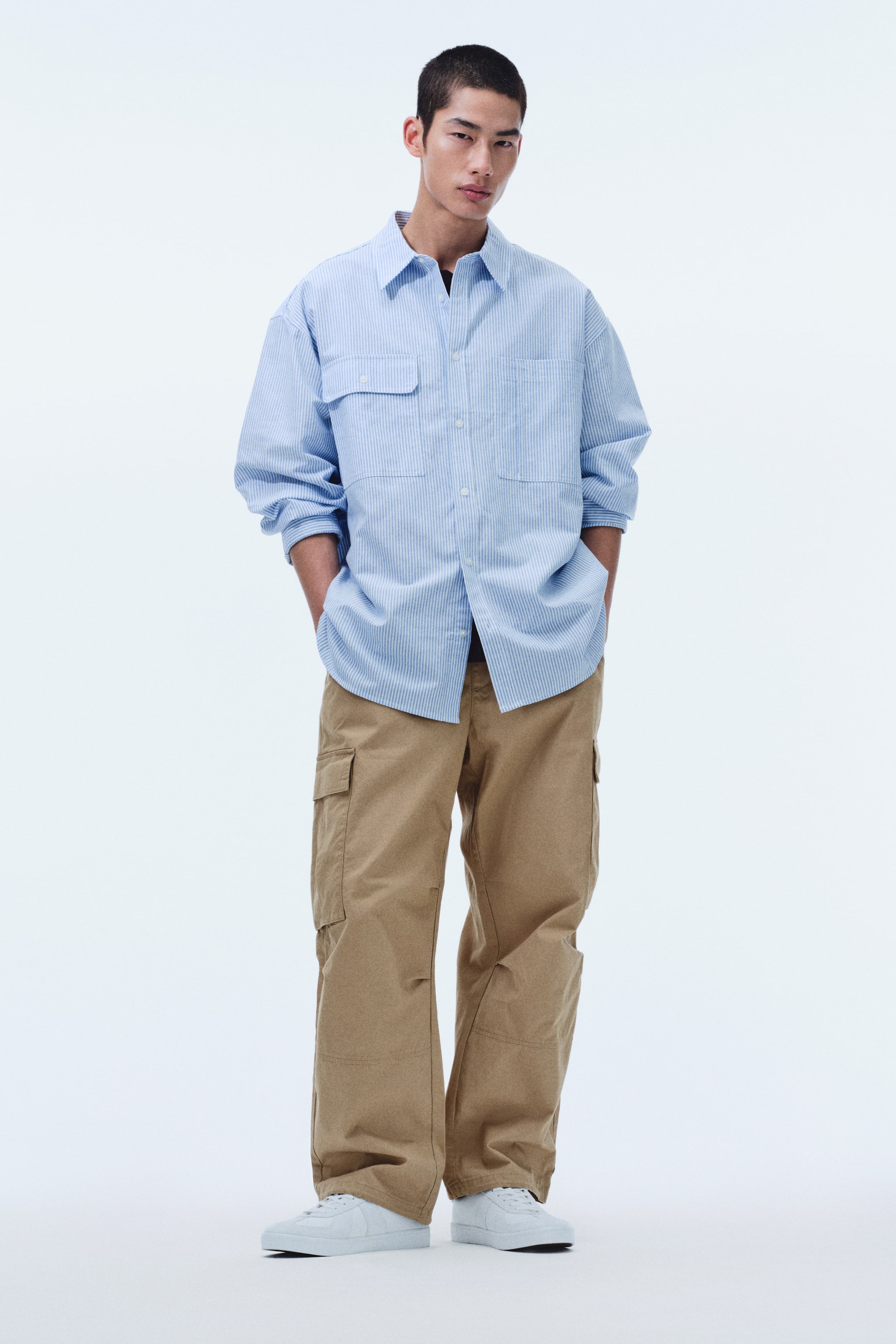 View larger image: A light blue and white striped long-sleeve shirt is shown in an oversized, relaxed fit from a front-facing view, featuring a classic spread collar and a full button-down placket with white buttons. It also displays two large patch pockets with buttoned flaps on the chest, and the long sleeves are pushed up, with the shirt worn open over a dark layer.
