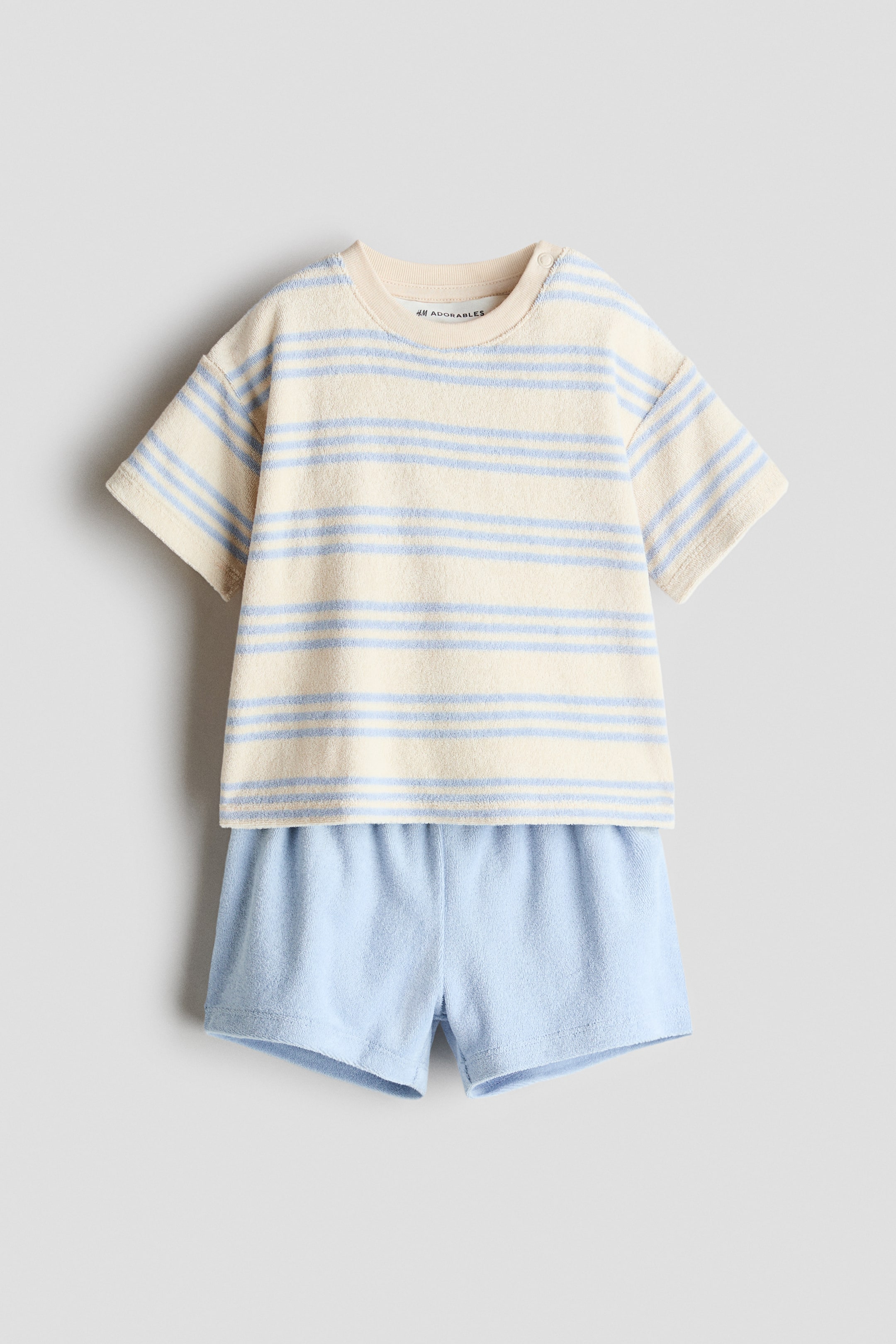Ver imagen más grande: Conjunto de bebé de dos piezas que incluye una camiseta de rizo de color crema con mangas cortas y rayas horizontales azul claro, un cuello redondo acanalado y un botón de presión visible en el hombro izquierdo. La camiseta se combina con unos pantalones cortos de rizo azul claro que presentan una cintura elástica.